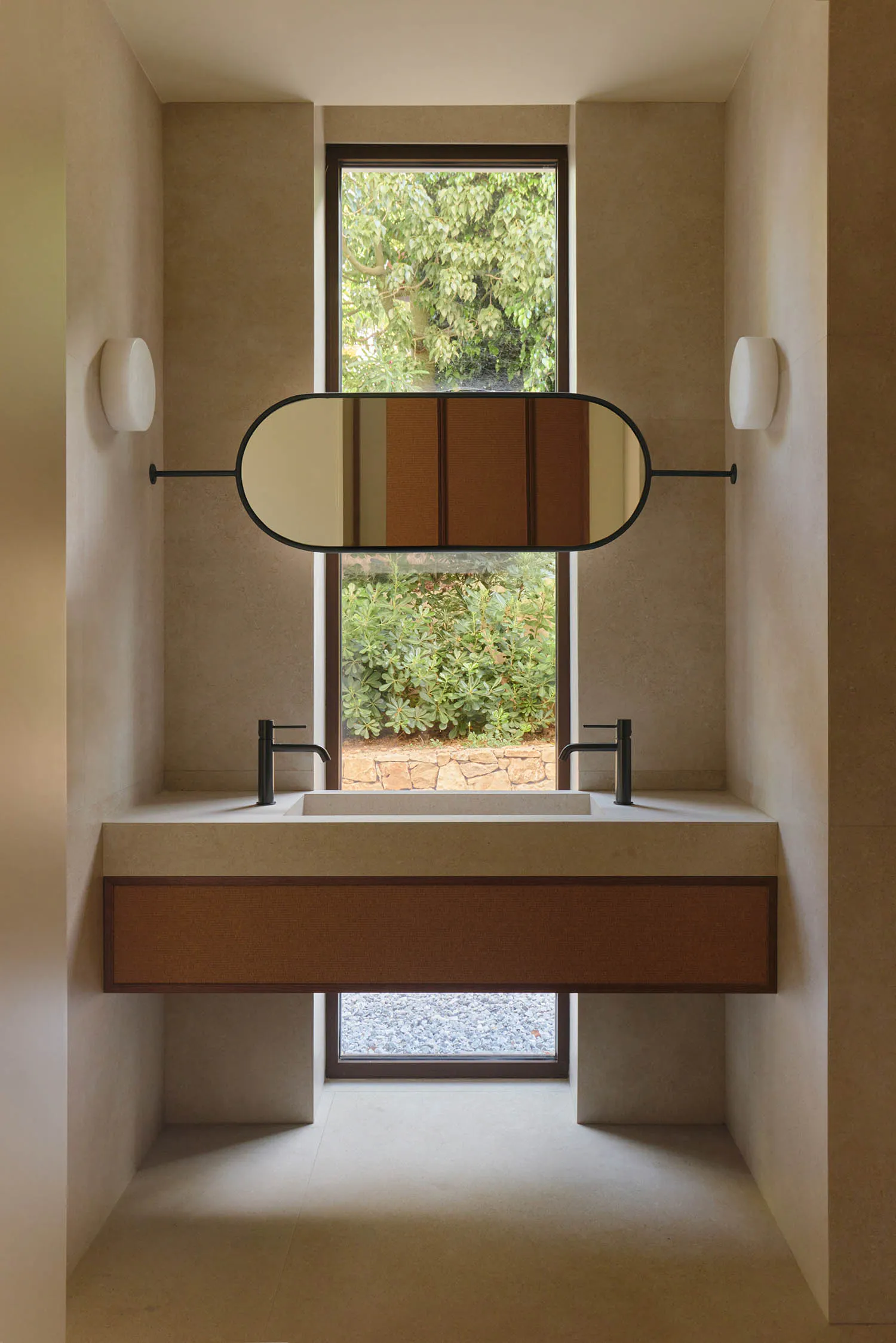 Modern minimalist bathroom with double concrete sink, black faucets, floating wooden vanity, oval mirror, and large windows overlooking a lush garden and stone pathway.