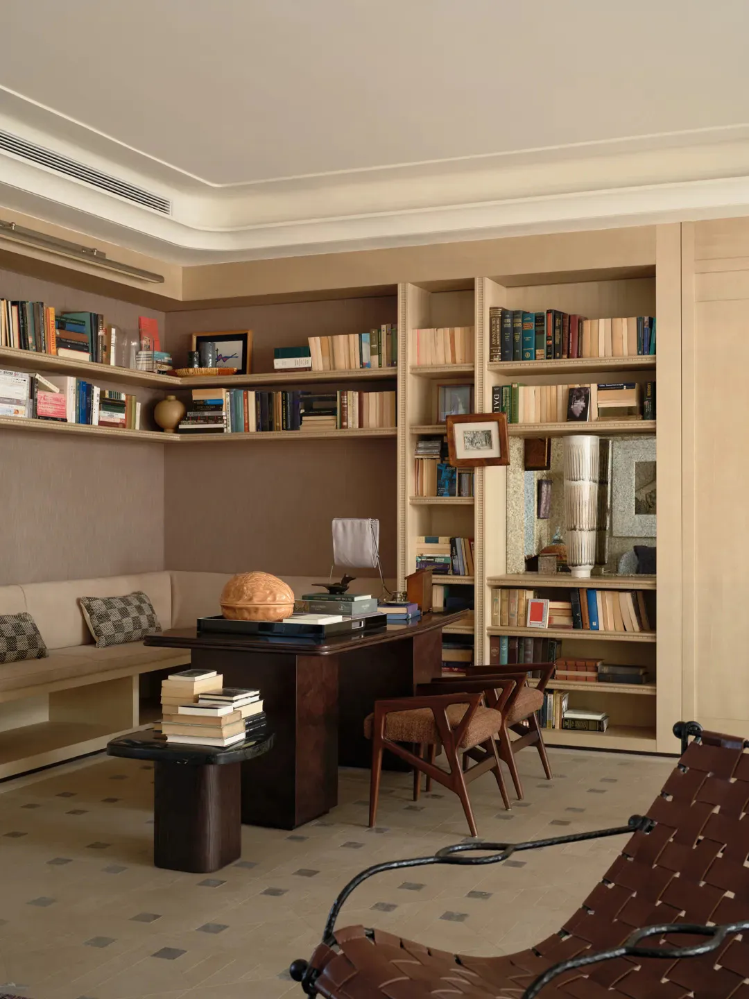 A warm and elegant home library featuring floor-to-ceiling built-in bookshelves, a large walnut desk, two mid-century wooden armchairs, a built-in window seat with checkered cushions, and a brown leather sling chair in the foreground.