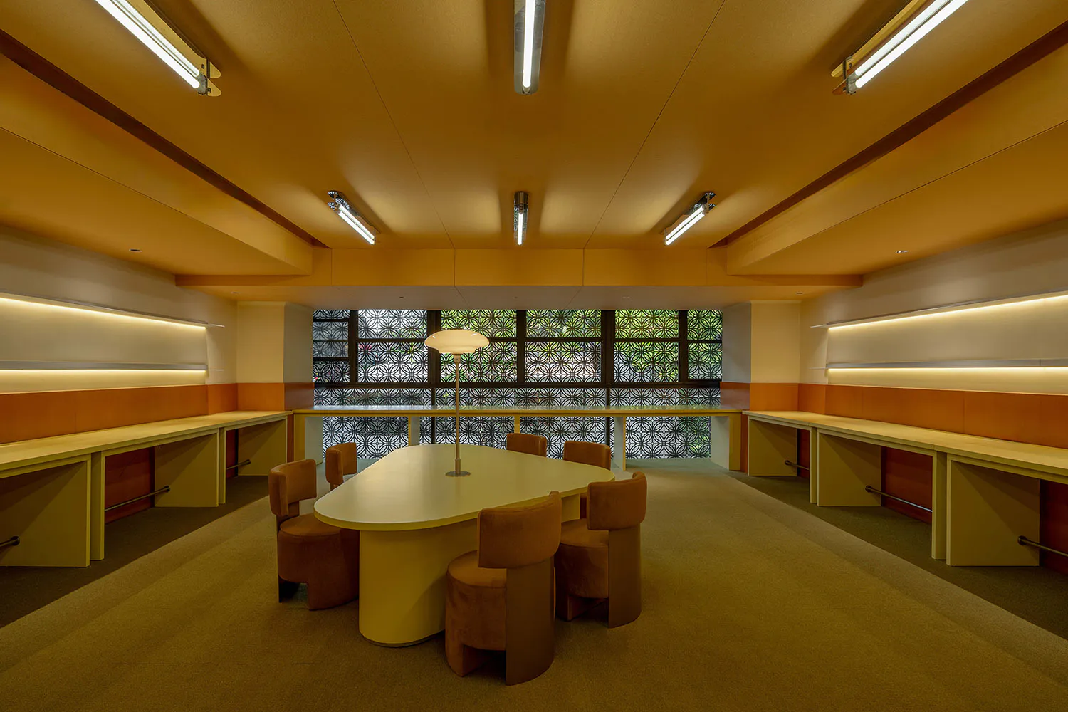 Modern institutional study room featuring a large oval cream table surrounded by six burnt-orange velvet swivel chairs, built-in light-wood perimeter desks with warm orange accent panels, and a large patterned window overlooking greenery. The ceiling has linear fluorescent lights and the carpet is a textured beige.