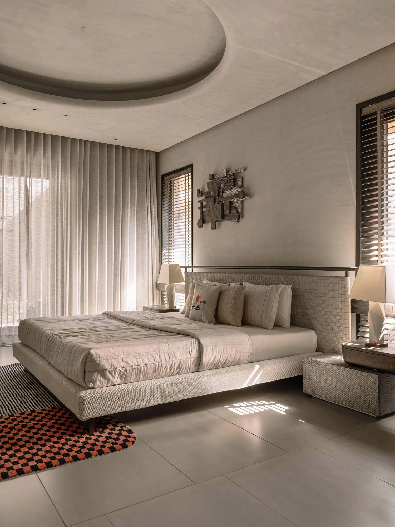 Modern minimalist bedroom featuring a curved concrete-like ceiling, textured plaster walls, a low platform bed with woven headboard, neutral bedding, two geometric table lamps, and colorful checkered and striped rugs.