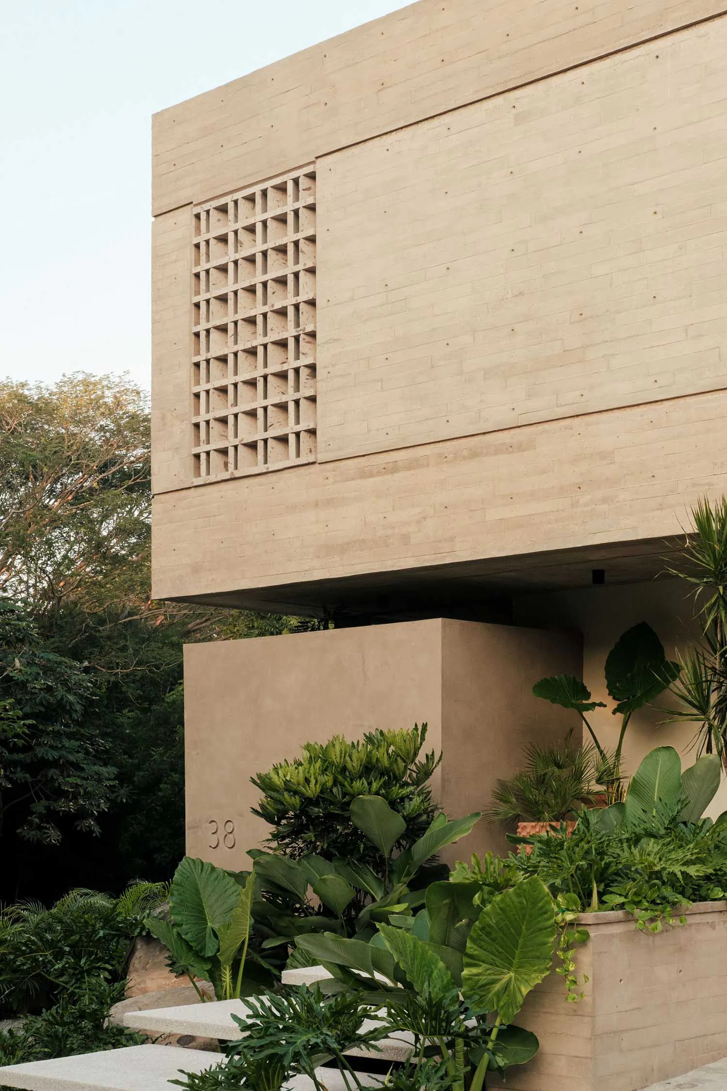 Exterior view of a modern concrete house with a decorative breeze-block wall and abundant tropical plants in the foreground.