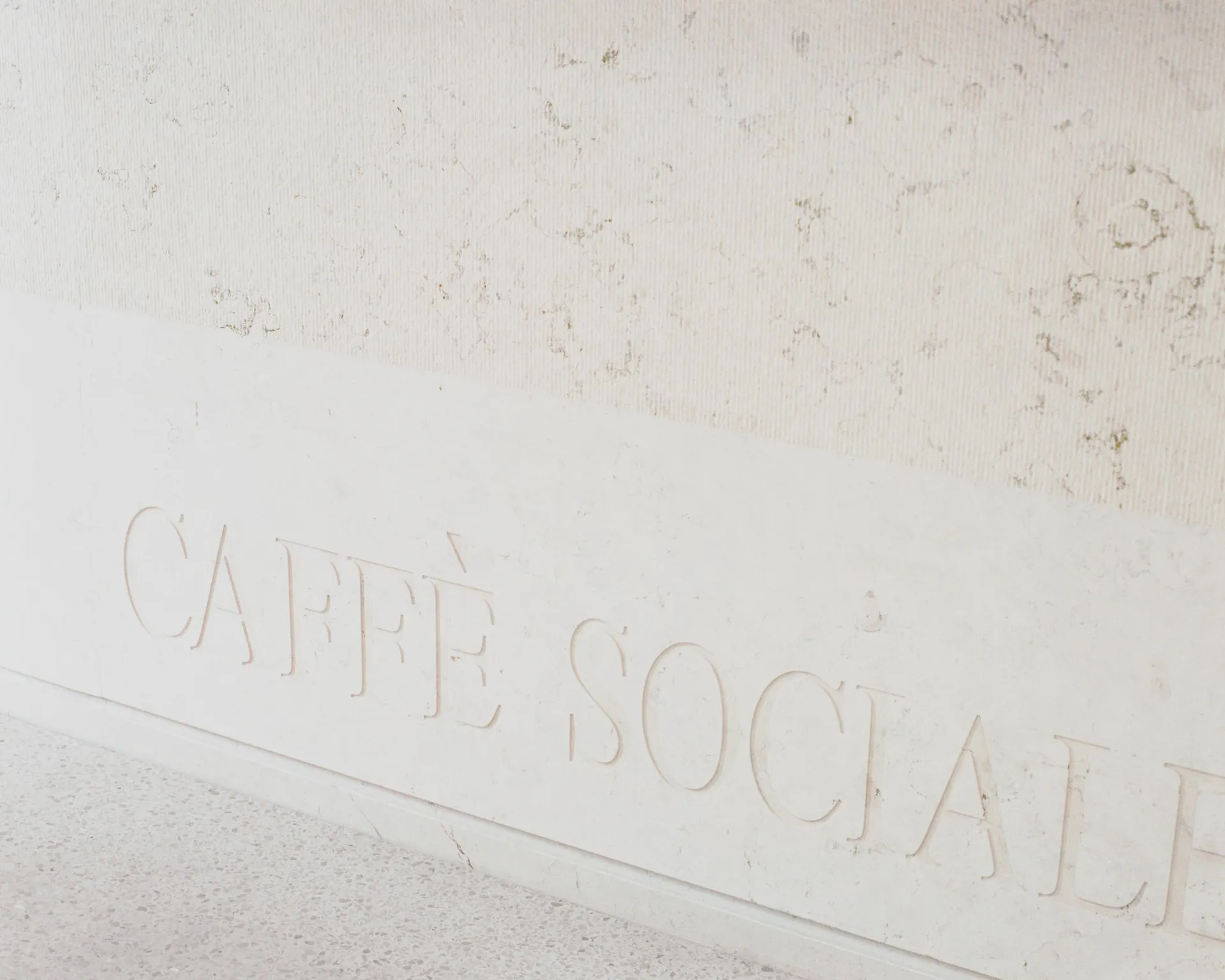 Close-up view of a white marble wall featuring raised, carved lettering that reads 'CAFFÈ SOCIALE' in an elegant serif font, with a speckled terrazzo floor visible below.