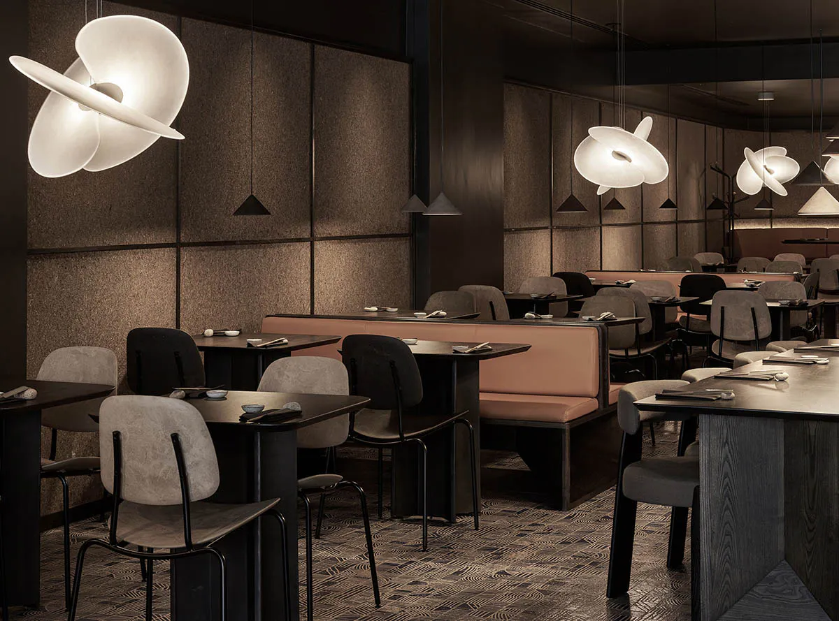 Modern Japanese restaurant interior with dark textured walls, warm wood tables, upholstered chairs, and large sculptural white pendant lights creating a moody elegant atmosphere.