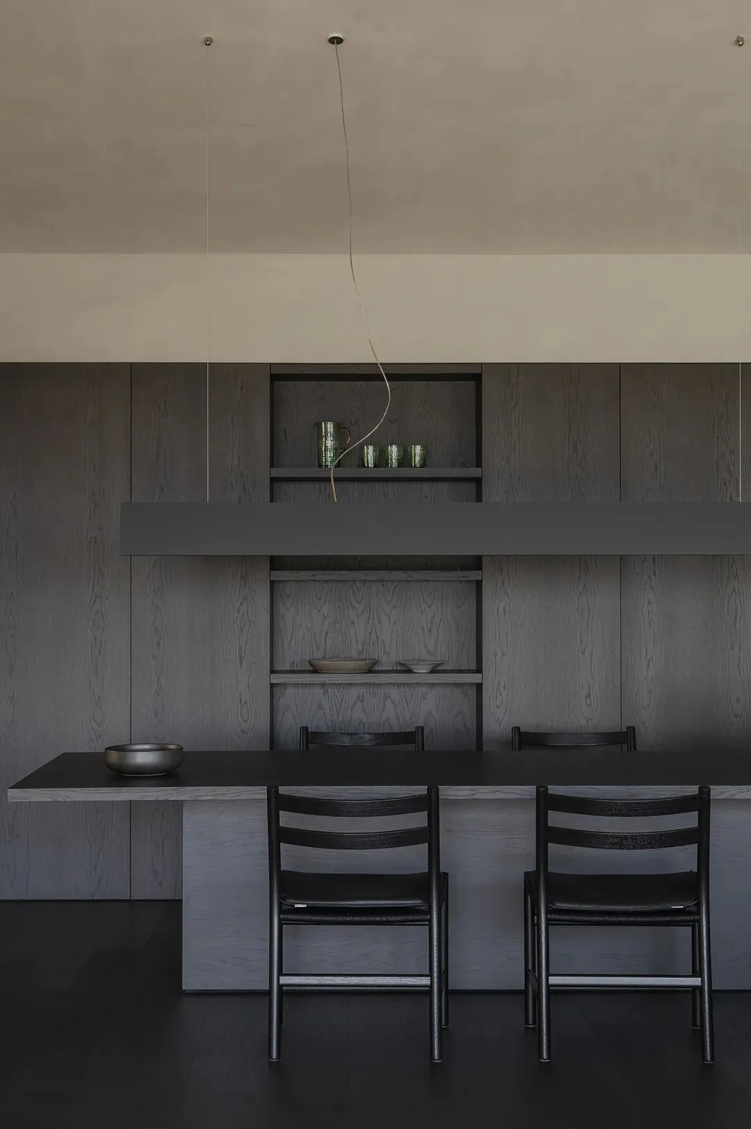 Modern minimalist dining room featuring dark wood cabinetry, floating open shelves, a long black dining table, and matching dark wood chairs under a single pendant light.