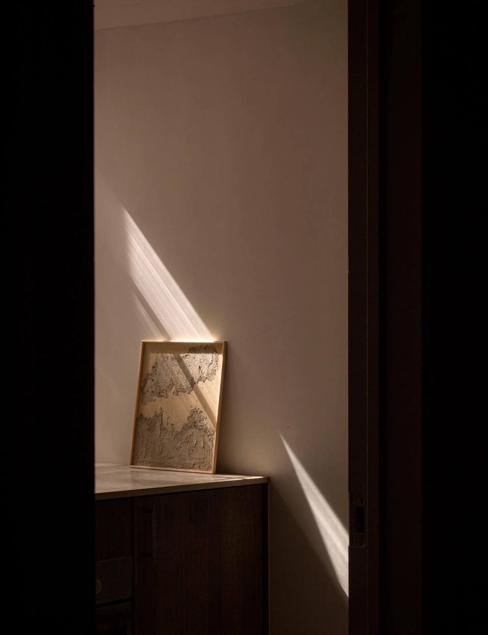 A framed textured artwork with an abstract, raised surface sits on a dark wood cabinet in a minimalist interior corner, dramatically lit by a diagonal shaft of sunlight casting strong shadows on the plain wall.