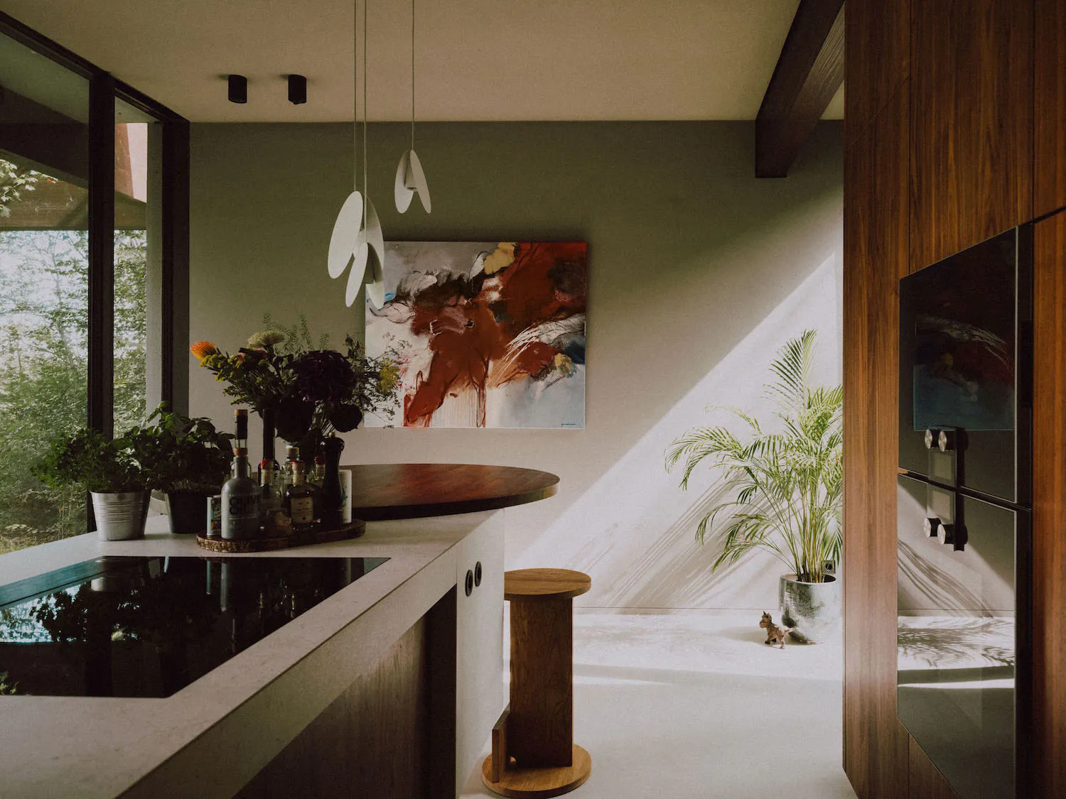 Modern kitchen interior with a curved walnut bar counter, black induction cooktop, pendant lights, large abstract painting, and potted plants in a bright, minimalist space with large windows.