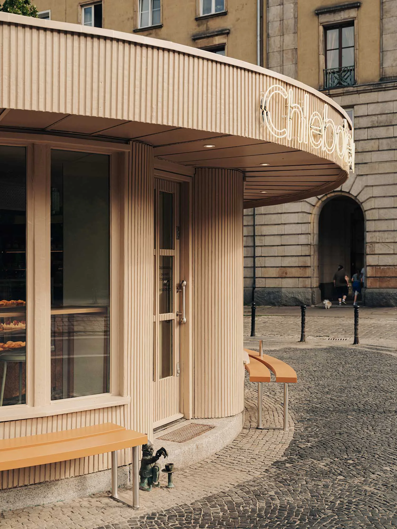Curved beige bakery storefront with vertical fluted cladding, glass display windows showing pastries, wooden benches, and a small bronze statue on a cobblestone square in Warsaw.