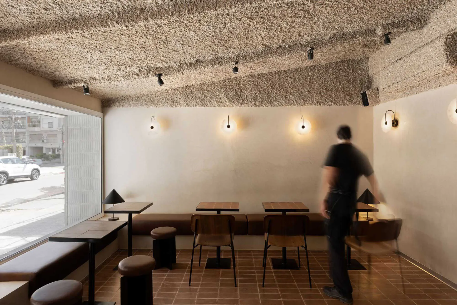 Interior view of a modern minimalist cafe with a heavily textured plaster ceiling, terracotta floor tiles, wooden tables, built-in banquette seating, and a person walking through the space.