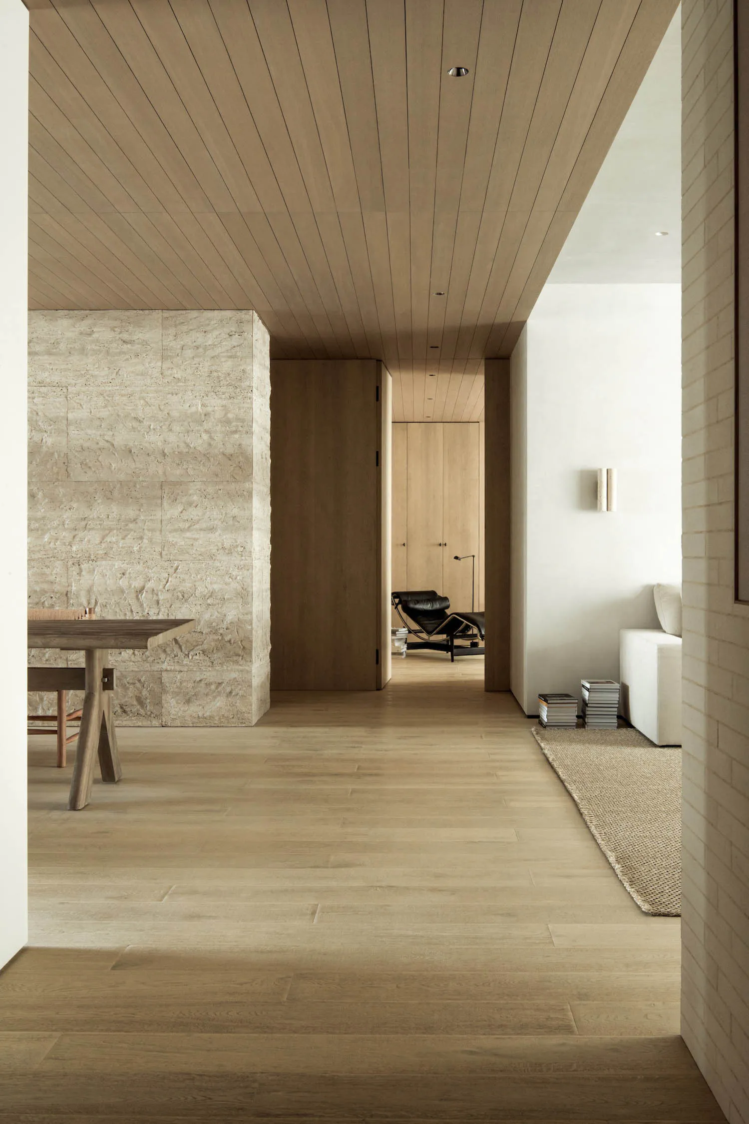 Modern minimalist interior hallway with a large textured travertine stone wall on the left, light oak wood plank ceiling and flooring, a wooden dining table, and a view into a living area with a black leather Eames lounge chair and white sofa.