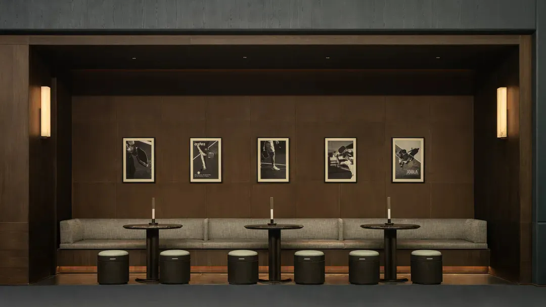 Modern lounge with dark wood paneled walls, linear wall sconces, upholstered bench seating, round black tables with candles, and framed black and white photographs of athletes including tennis, baseball, and hockey players.