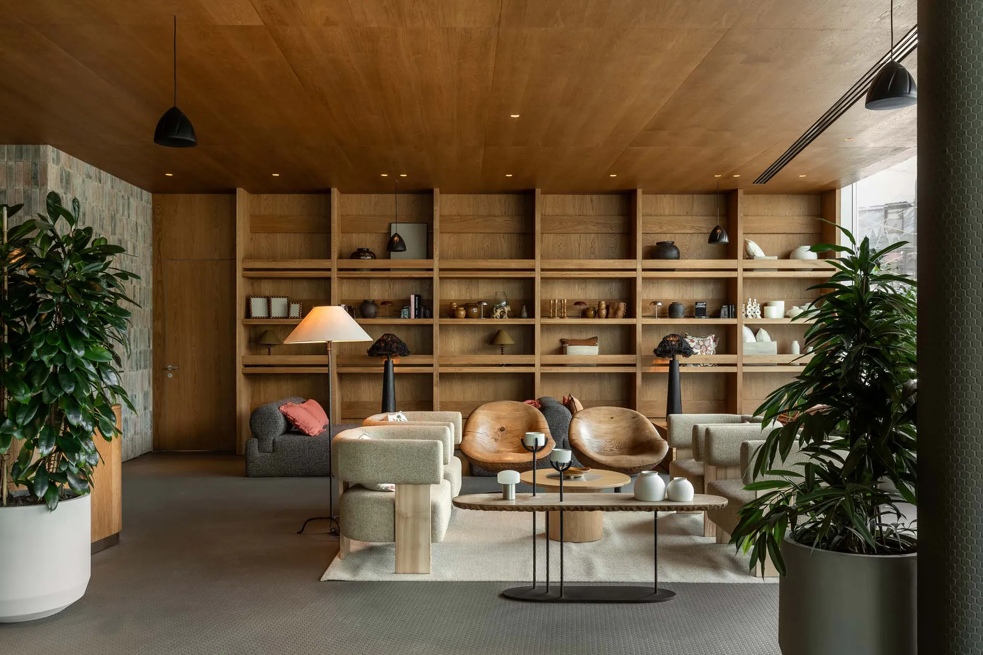 Modern living room featuring extensive oak built-in shelving, sculptural wooden lounge chairs, textured fabric sofas, large potted plants, and layered lighting under a wood-paneled ceiling.
