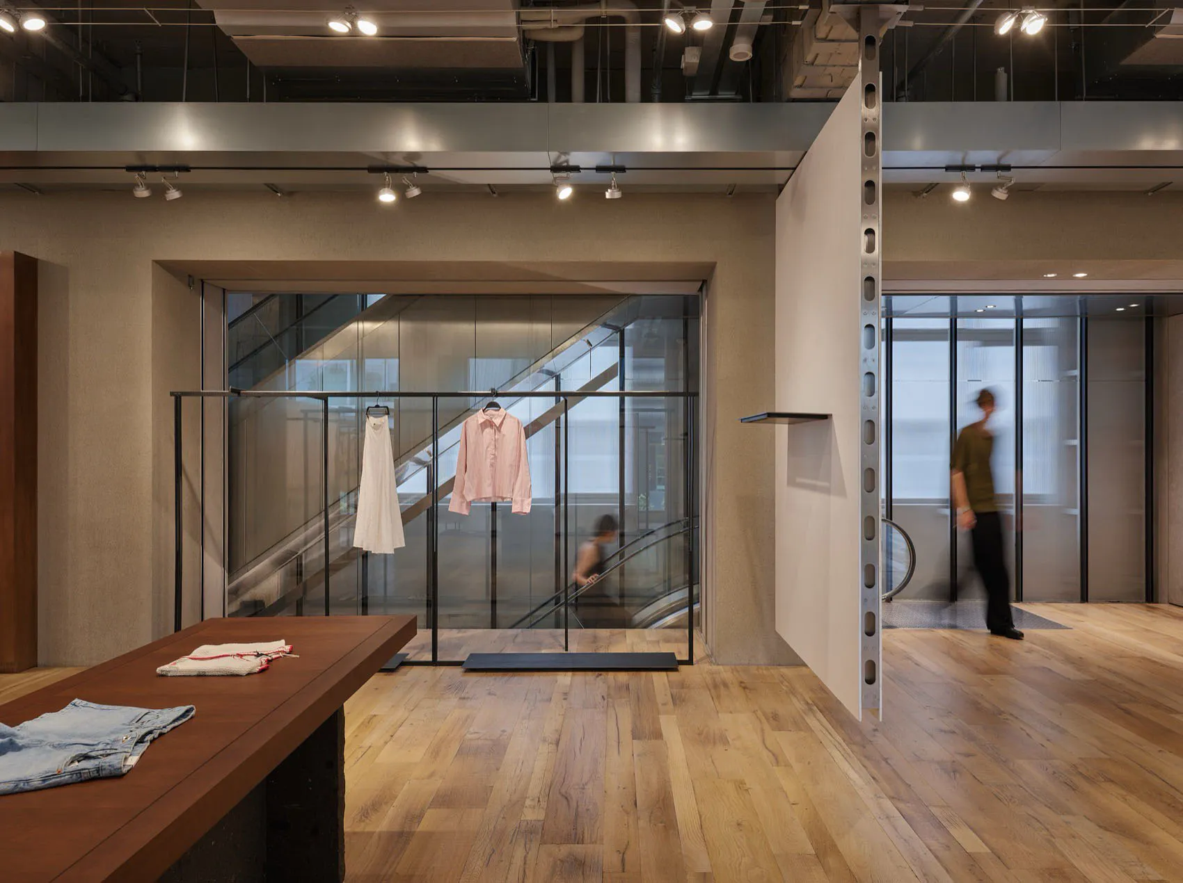 現代極簡服裝精品店室內空間,淺色橡木地板、裸露鋼梁、軌道燈,以及在玻璃樓梯背景前以工業衣架展示的服裝。