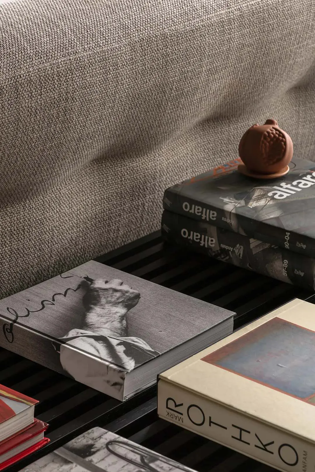 Close-up view of a textured light gray linen sofa with a black slatted coffee table holding multiple art books including titles by Alfaro and a terracotta pomegranate-shaped sculpture on top.