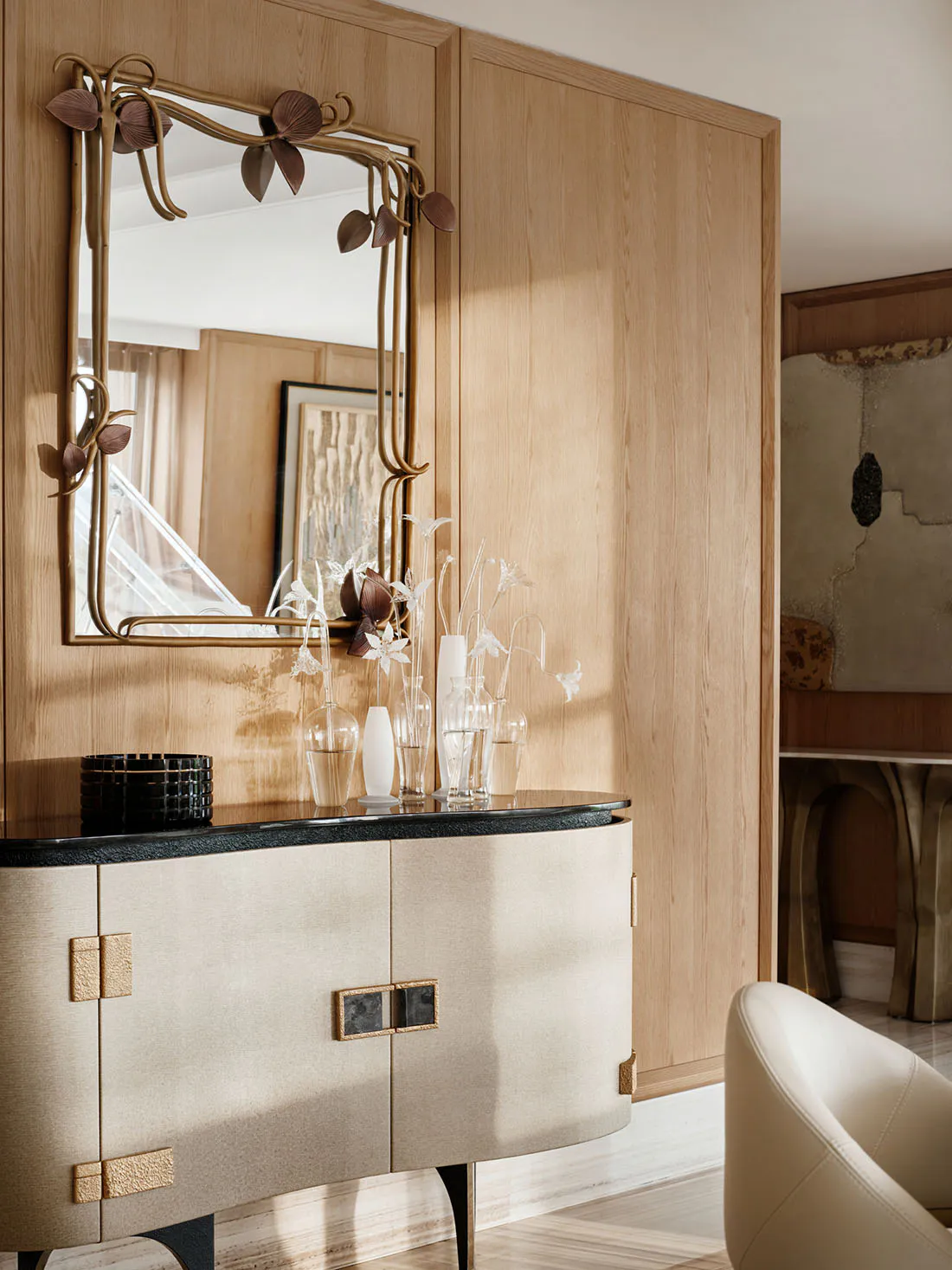 Elegant entryway featuring a curved beige console table with black marble top, paired with an ornate brass mirror decorated with botanical motifs, set against light oak wood paneling.