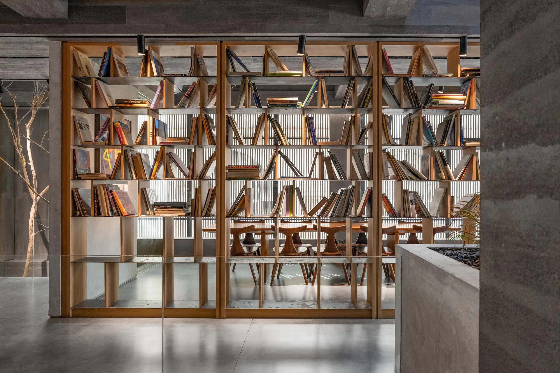 Modern concrete interior featuring a large double-sided oak bookshelf filled with books, wooden reading tables and stools visible through the shelves, with concrete walls and polished floor.