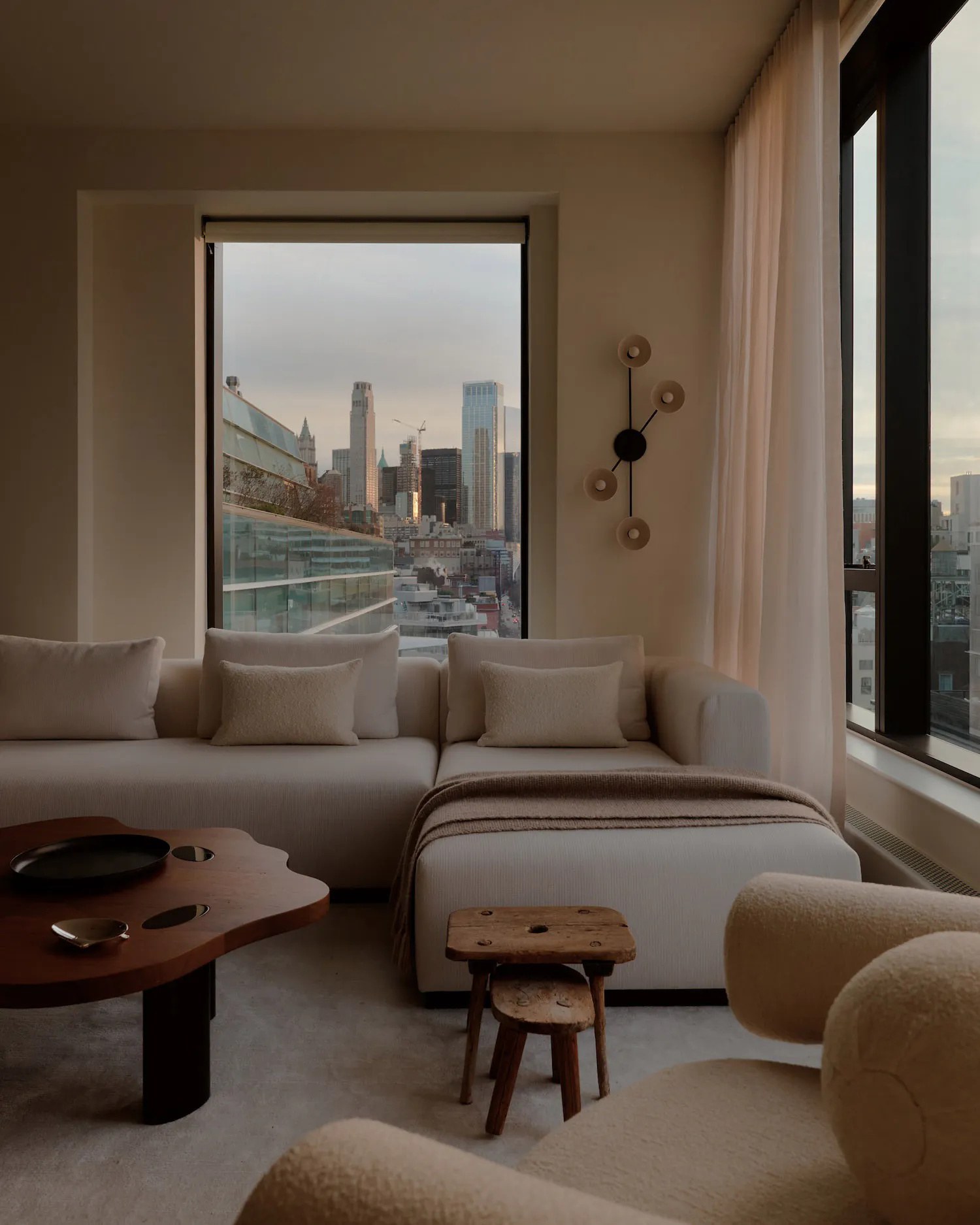 Modern minimalist living room featuring a large L-shaped boucle sofa, wooden coffee table, and large windows overlooking a city skyline at dusk.