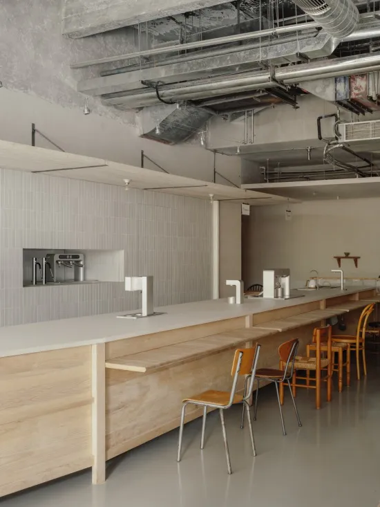 Modern minimalist cafe interior with a long light-wood plywood counter, metal stools with orange seats, exposed silver HVAC ducts on a concrete ceiling, and a recessed stainless steel coffee station.