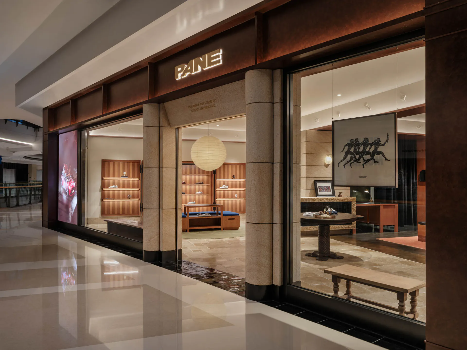 Exterior view of a luxury shoe store named PANE with large glass windows, travertine stone pillars, wooden shelving, and a hanging paper lantern inside.