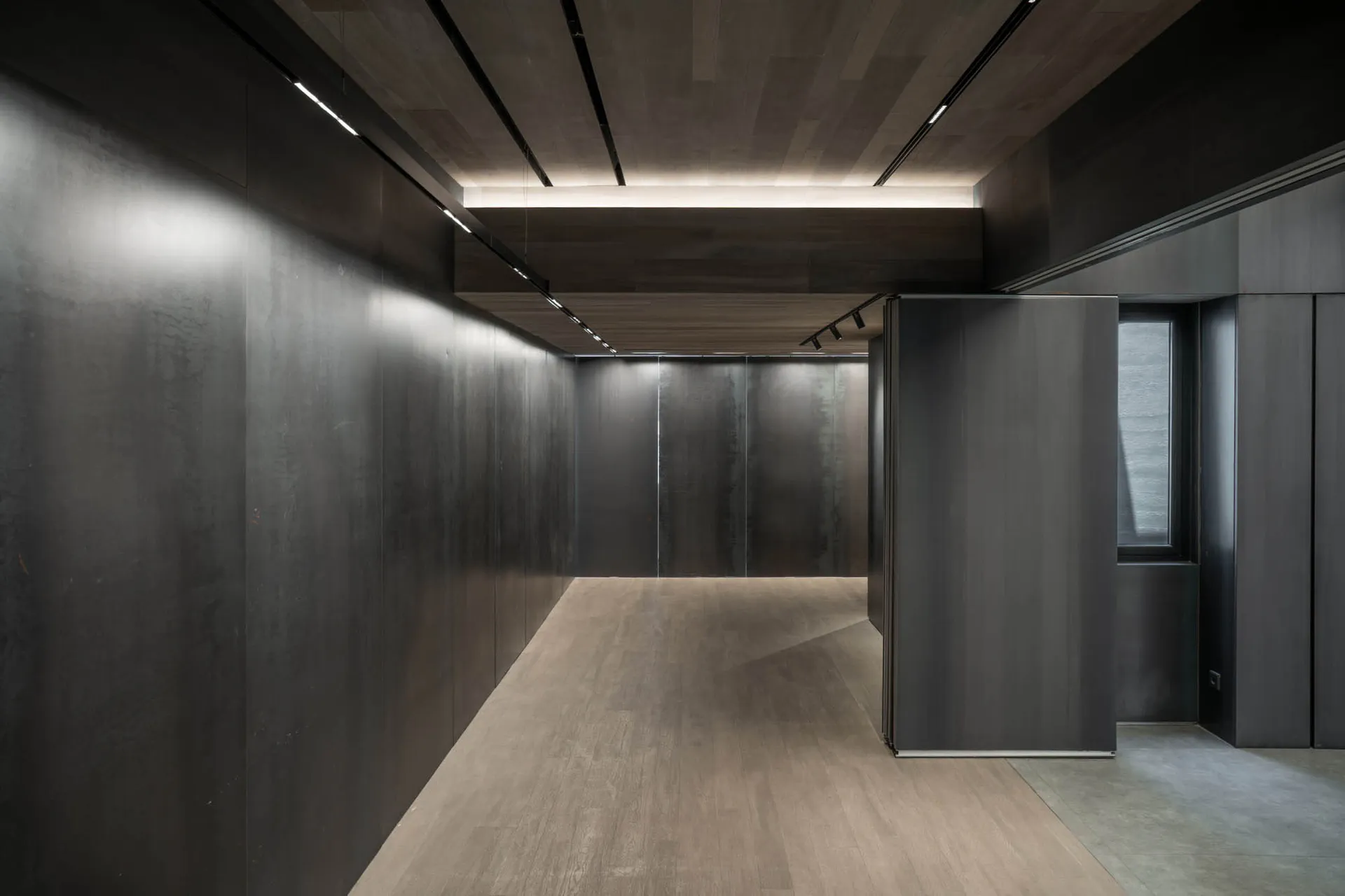 Modern minimalist corridor with dark metal or concrete panel walls, light wood flooring, and recessed linear ceiling lights creating a dramatic atmosphere.