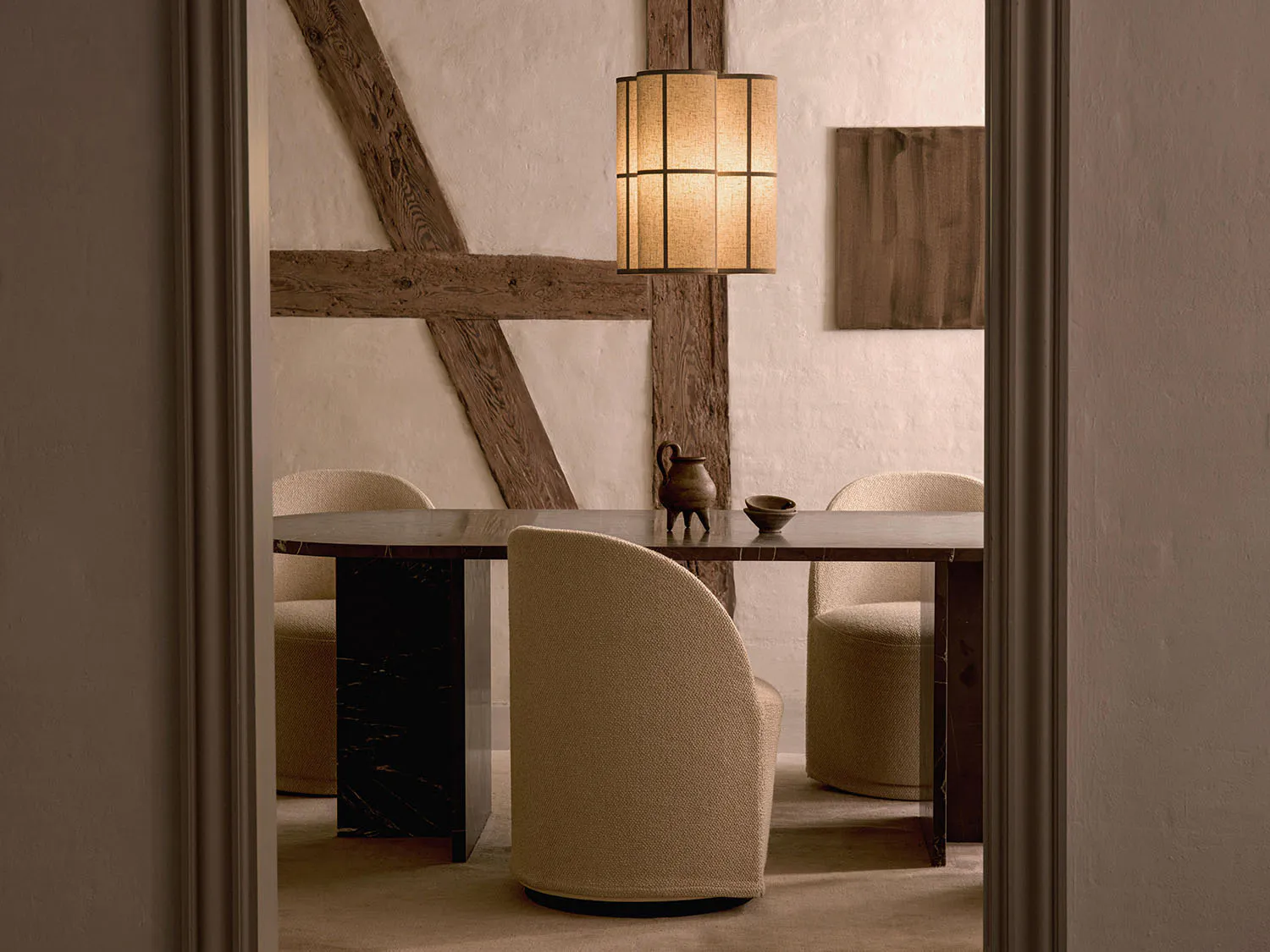 View into a minimalist dining room with a dark marble dining table, four cream boucle swivel chairs, a ceramic pitcher and bowl centerpiece, exposed wooden beams on white plaster walls, and a cylindrical woven pendant lamp.