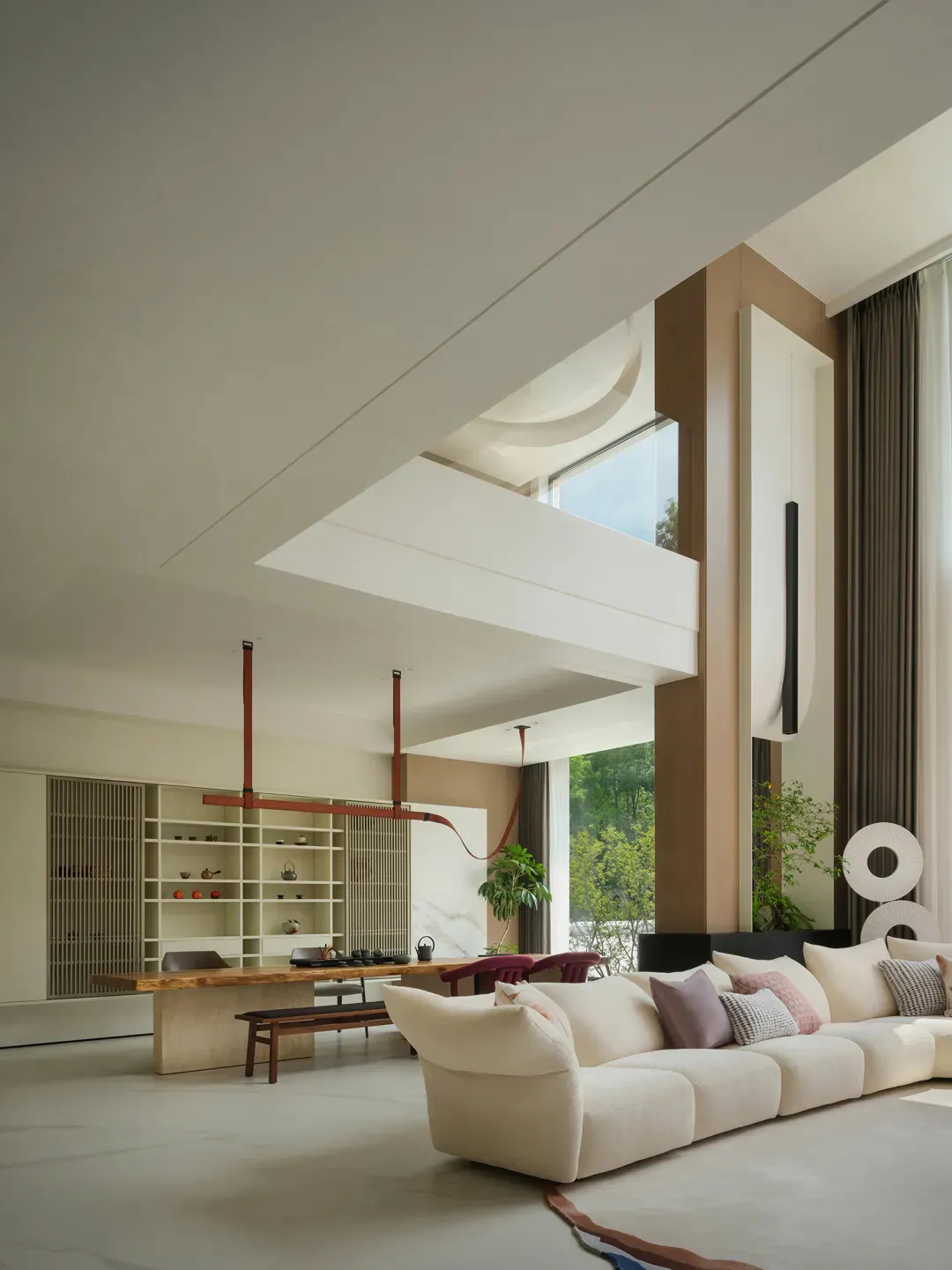 Modern minimalist living room with double-height ceiling, large cream sectional sofa, live-edge oak dining table, and warm neutral walls with large windows overlooking greenery.