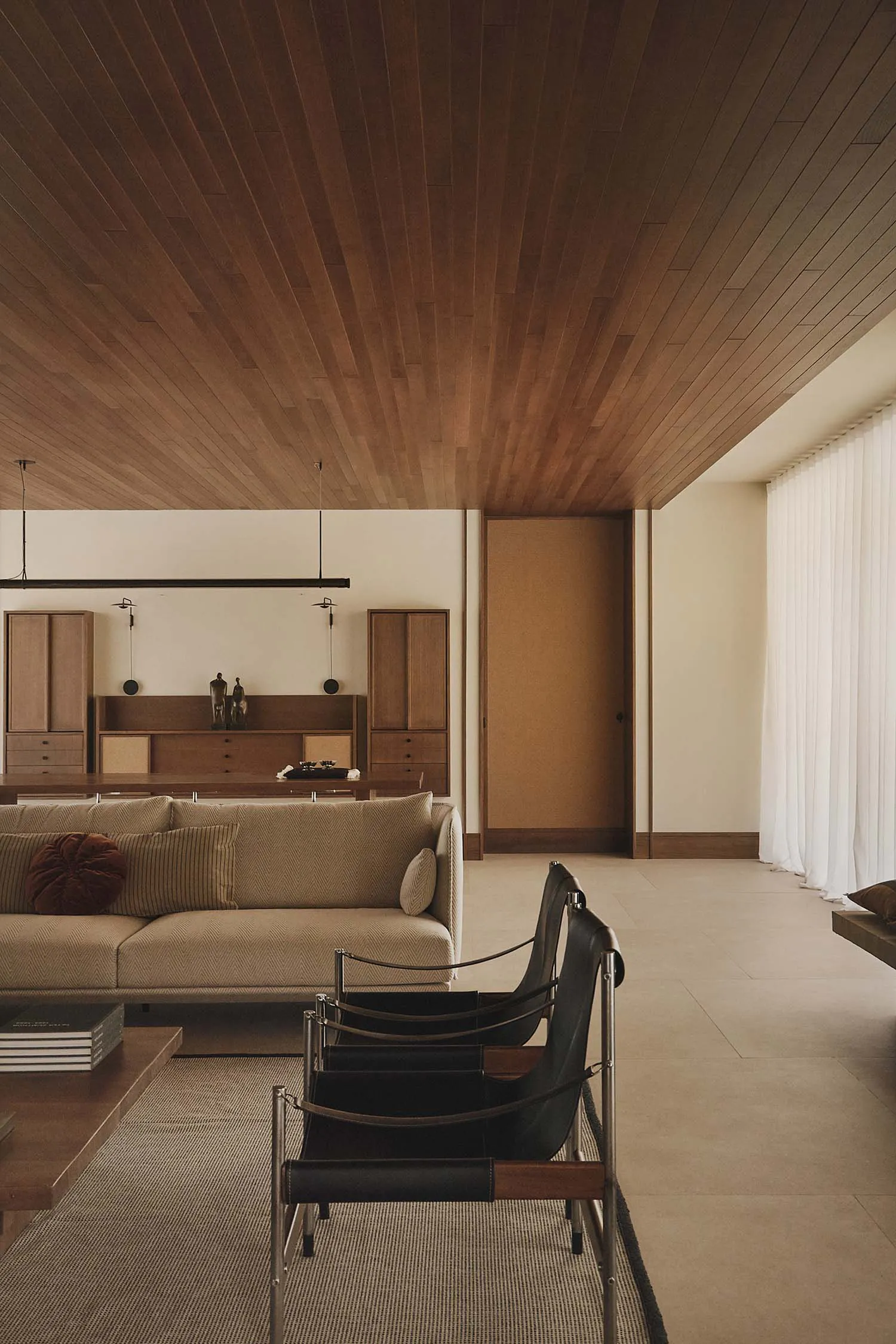 Modern minimalist living room featuring a dramatic wood-paneled ceiling, a beige herringbone sofa with rust and striped cushions, two black leather and chrome sling chairs in the foreground, and built-in wooden cabinetry against a white wall.