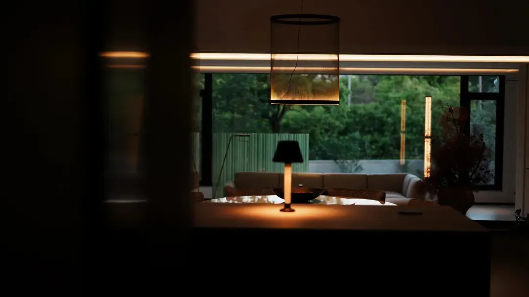 Modern living room interior featuring a large cylindrical pendant lamp, wooden dining table with lit table lamp, neutral sofa, and large windows overlooking a lush green garden at dusk.