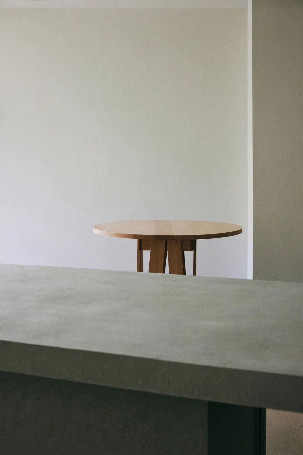 A round solid oak dining table with a sculptural pedestal base positioned in a minimalist room with concrete surfaces and neutral walls.