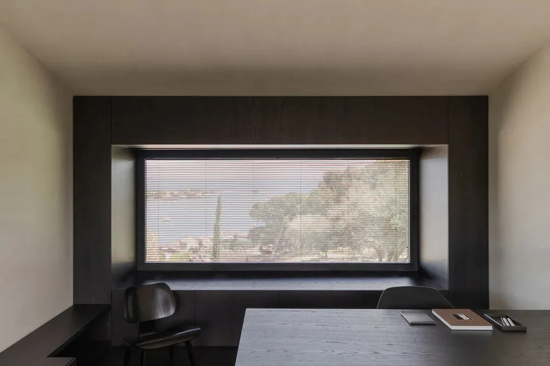 Minimalist home office interior featuring a large horizontal window with blinds, a built-in dark wood window seat, a black molded chair, and a concrete-like desk with minimal accessories.