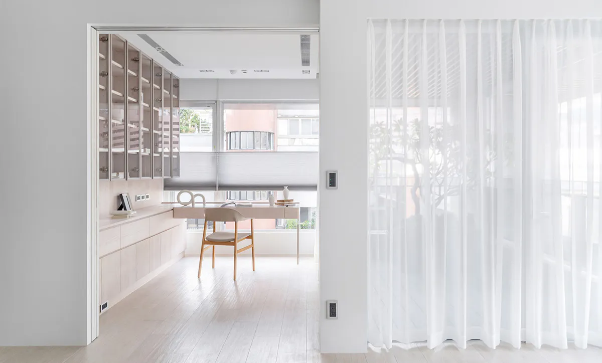 View into a bright minimalist home office featuring light wood built-in cabinetry, a simple wooden desk and chair, large windows with roller blinds, and white sheer curtains on the right.