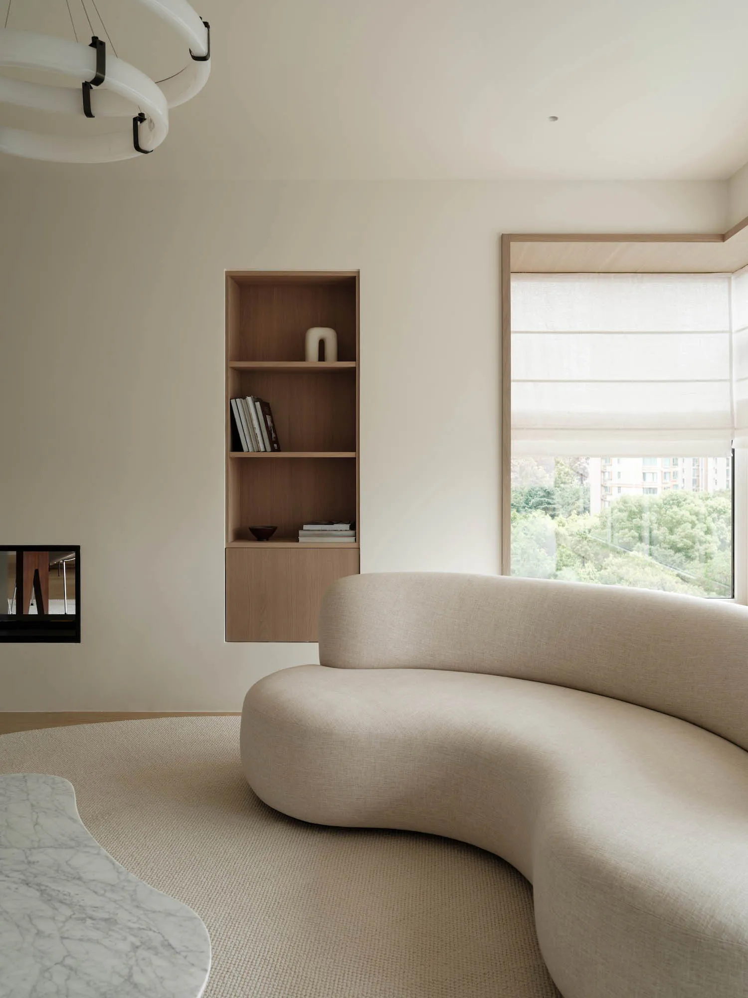 Modern minimalist living room featuring a large curved beige boucle sofa, built-in oak bookshelf, marble coffee table, and large window with natural daylight.