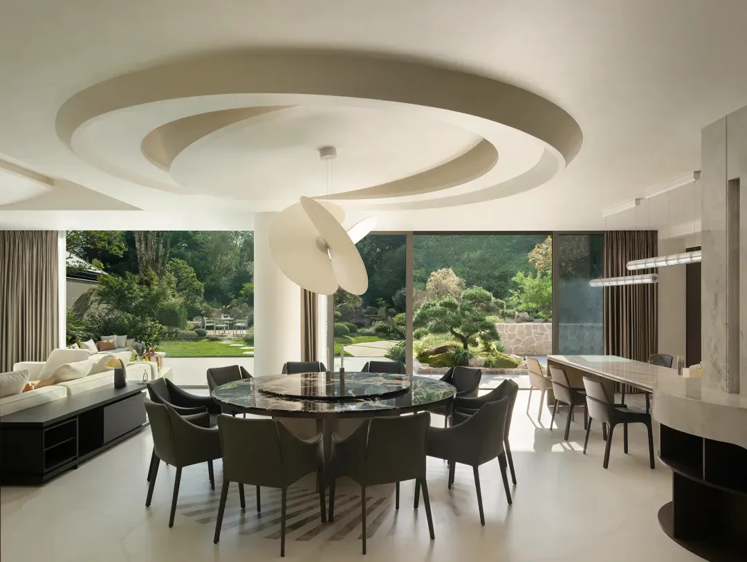 Modern luxury dining room with a large round dark marble table surrounded by eight dark green upholstered chairs, a sculptural white pendant light hanging from a circular coffered ceiling, and large glass doors opening to a lush Japanese-inspired garden.