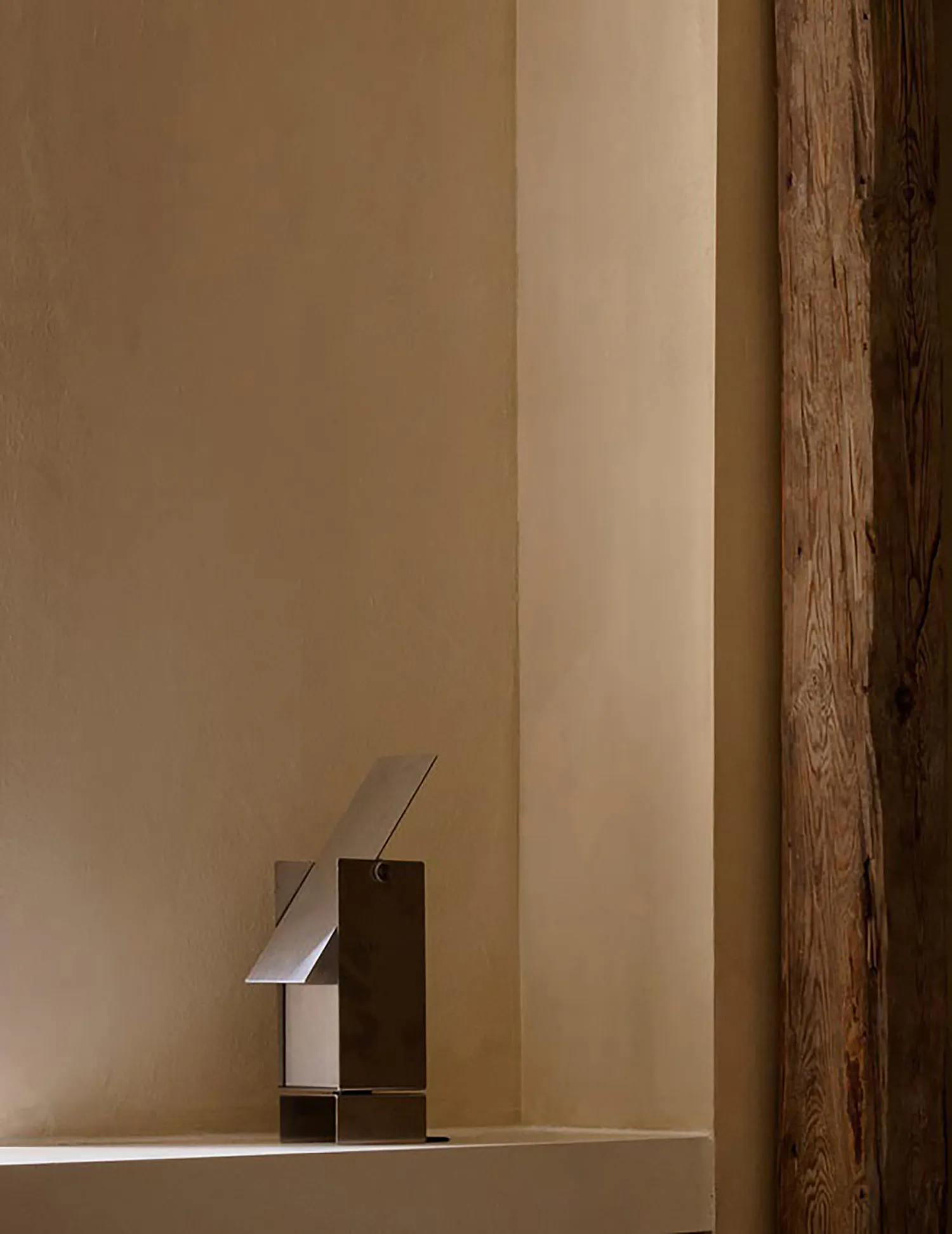 Modern stainless steel abstract sculpture placed on a white plaster windowsill next to a textured wooden beam in a minimalist interior space.