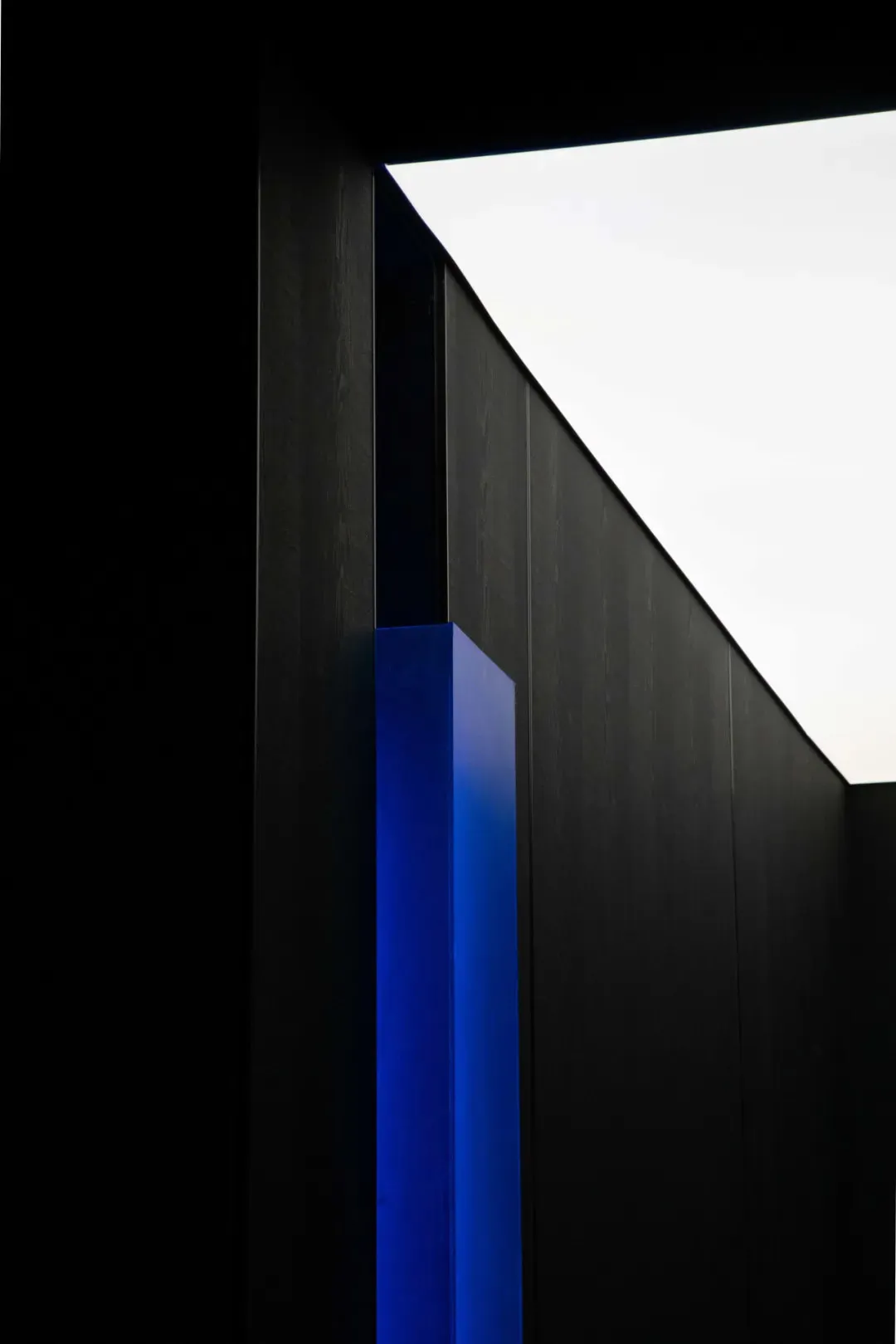 Low-angle view of a dark wooden architectural wall with a glowing bright blue vertical resin or acrylic column against an overcast sky.