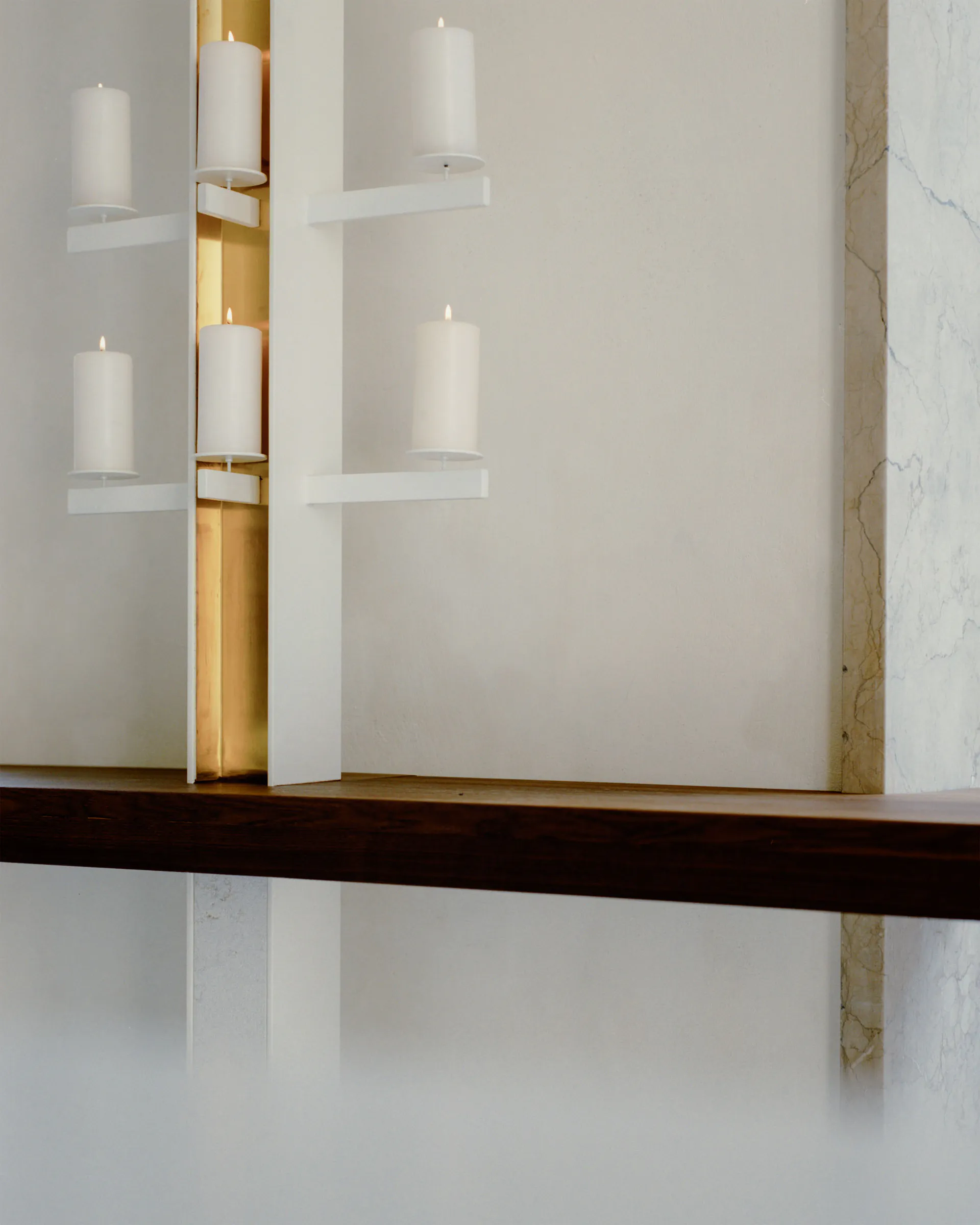 Modern minimalist wall installation with white pillar candles on white floating shelves, set against a recessed brass panel with warm backlighting, above a dark wood mantel and next to a white marble wall.