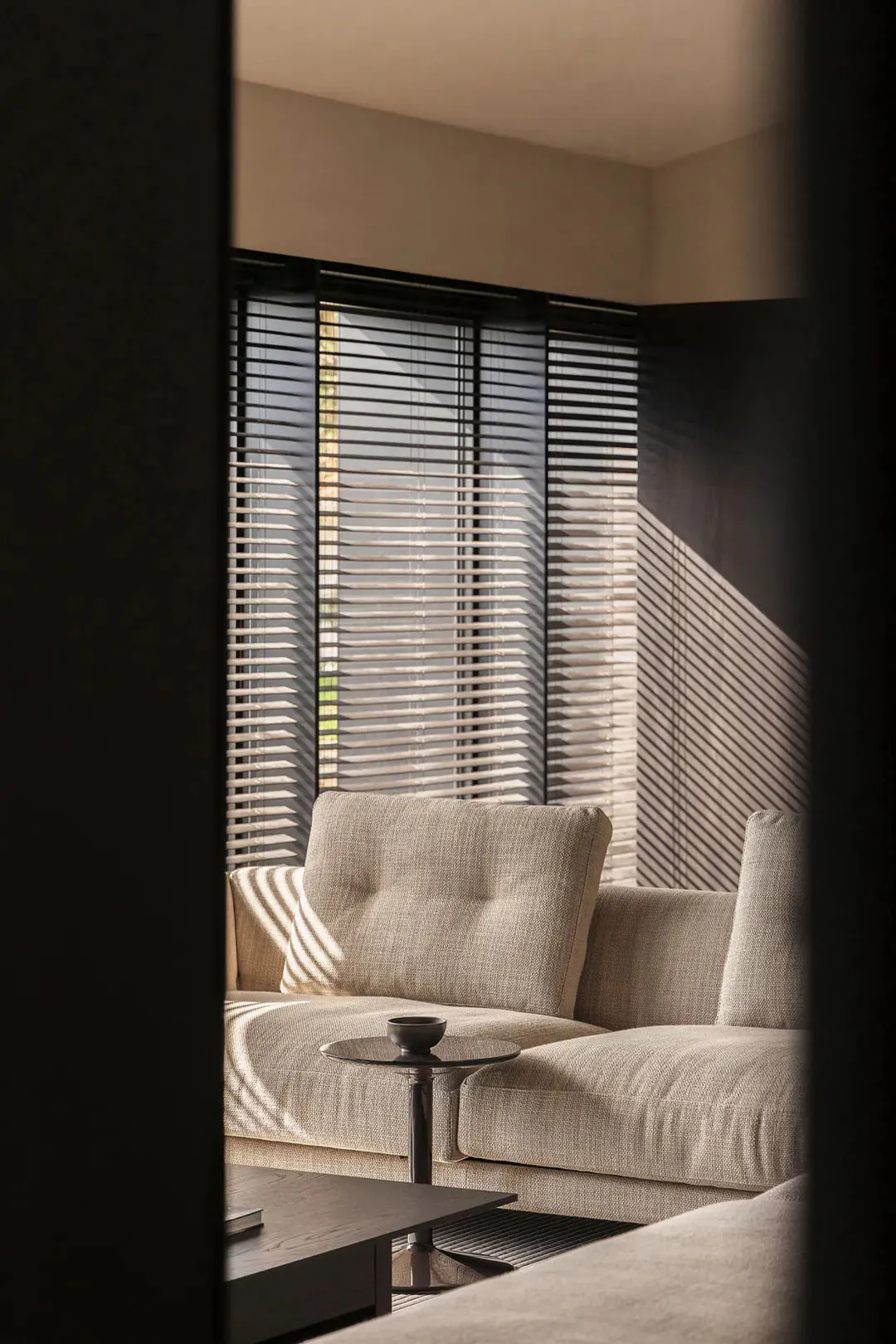 A contemporary living room corner featuring a beige boucle fabric sofa, round black side table with a small bowl, dark wood coffee table, and large windows with horizontal blinds casting striped shadows on the wall and furniture.