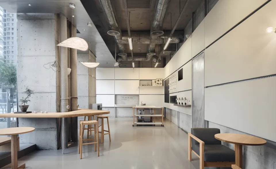 Modern minimalist cafe interior featuring exposed concrete walls, a long curved wooden counter with stools, white cabinetry, and industrial ductwork visible on the ceiling.