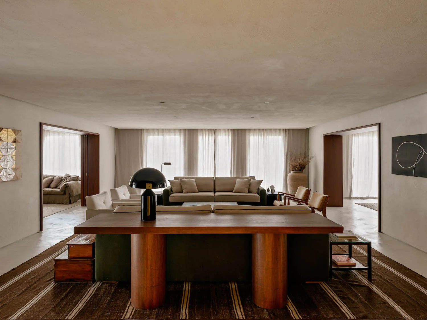 A spacious modern minimalist living room featuring a large round walnut dining table in the foreground, a green sectional sofa, a beige sofa in the background, and large windows with sheer curtains allowing soft daylight to fill the space.
