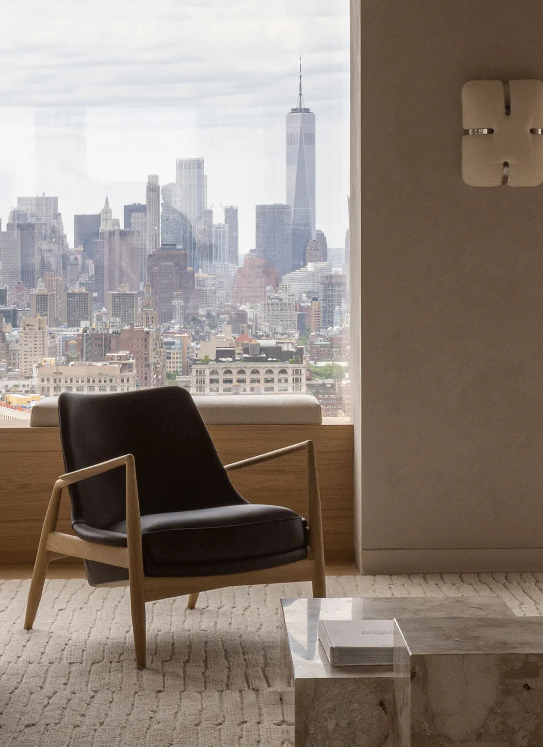 Modern oak and black leather armchair placed on a textured rug in front of a large window overlooking the New York City skyline with One World Trade Center prominent, next to a marble coffee table and a modern wall sconce.