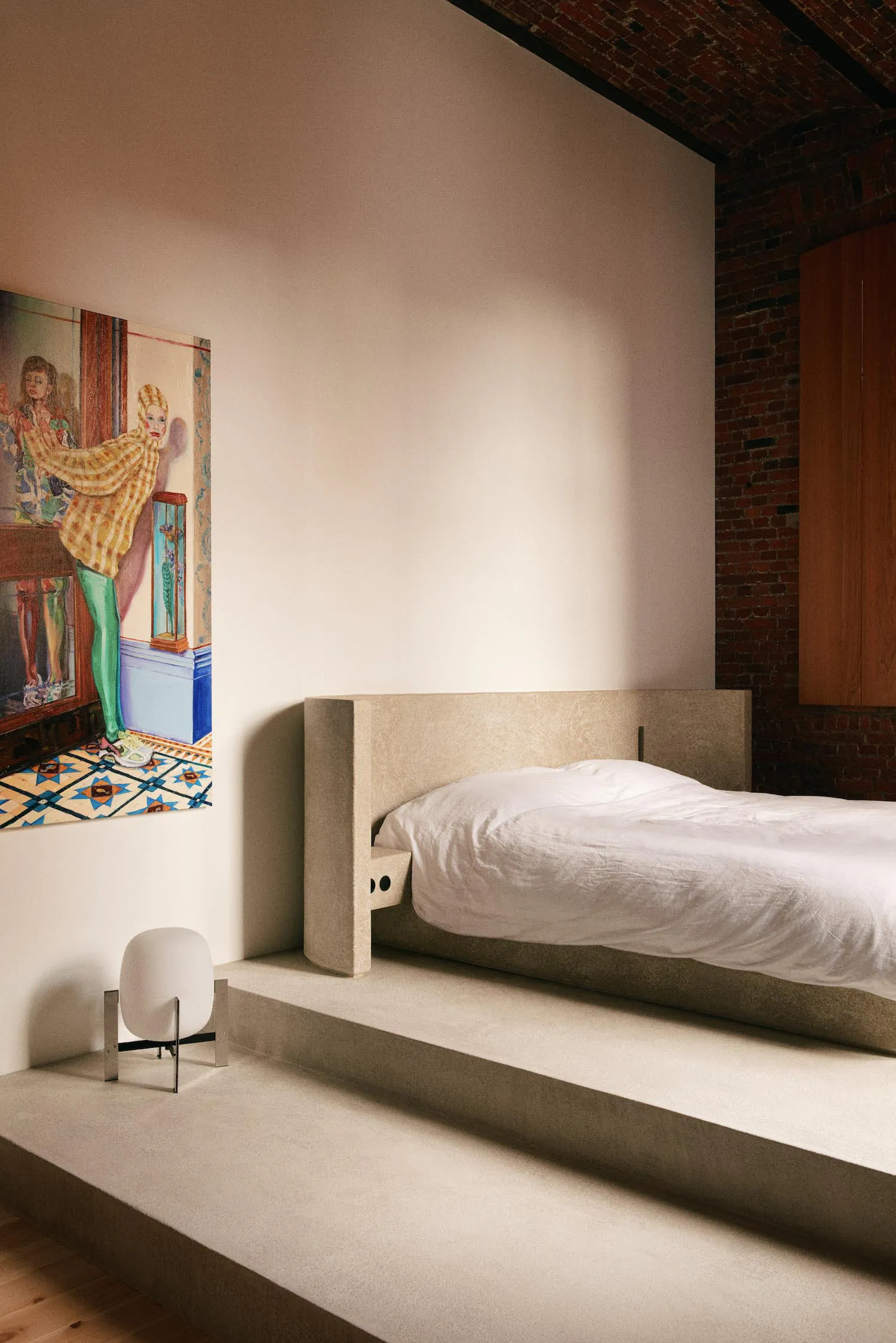 Modern minimalist bedroom with a low concrete platform bed dressed in white linens, a large colorful figurative painting on the wall, a white mushroom lamp on a stepped concrete platform, and exposed red brick wall in the background.