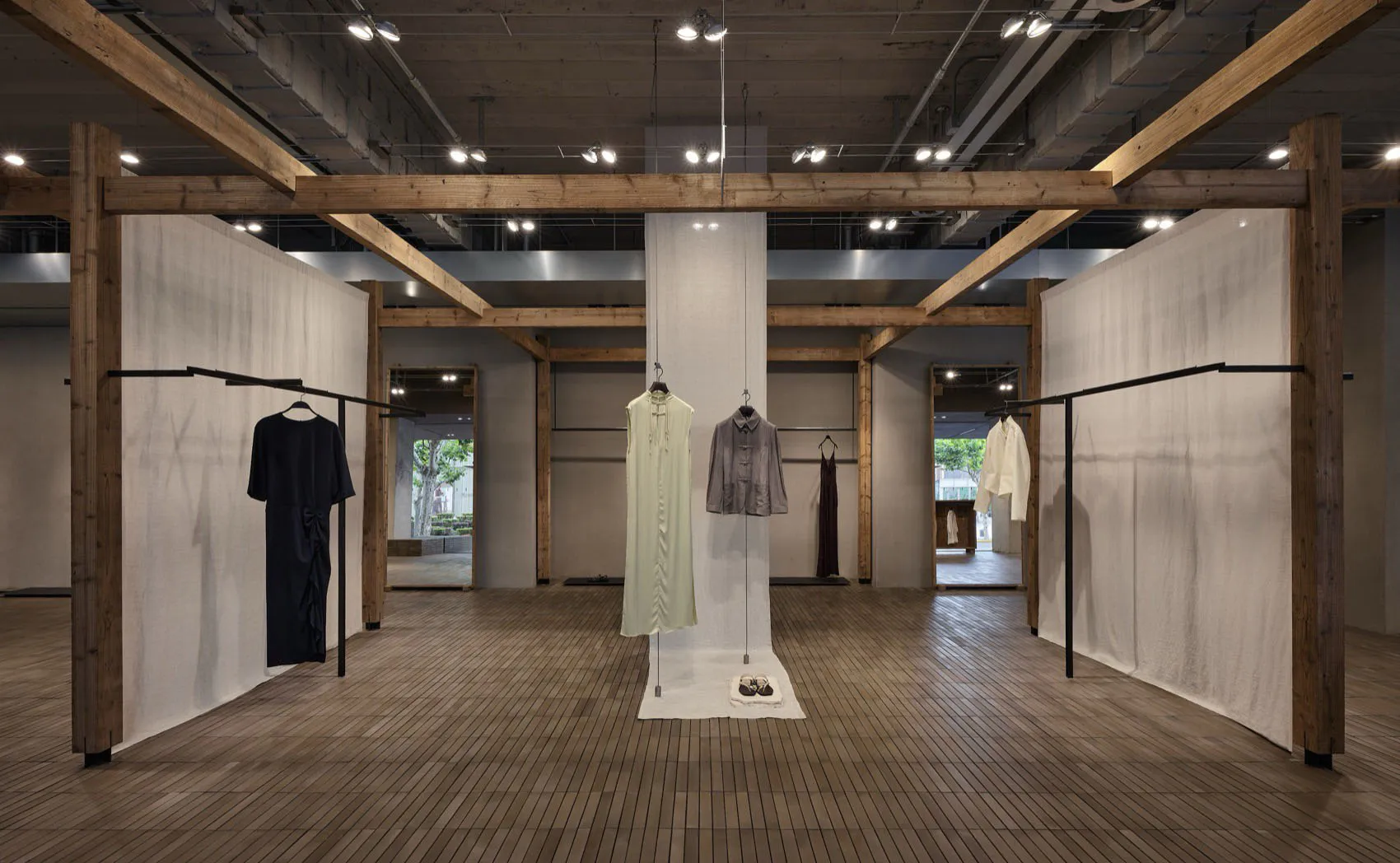 寬敞的極簡快閃店內部,展現裸露木結構樑柱、混凝土牆面、淺色木地板,以及懸掛在黑色衣架與中央展示桿上的設計師服裝。