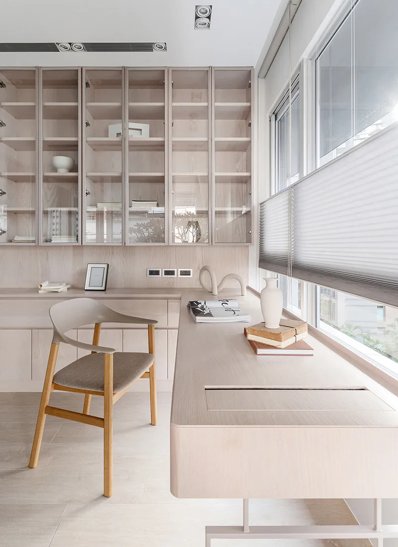 Modern minimalist home office with light wood built-in cabinetry, a wooden chair with upholstered seat, large windows with blinds, and decorative objects on the desk.