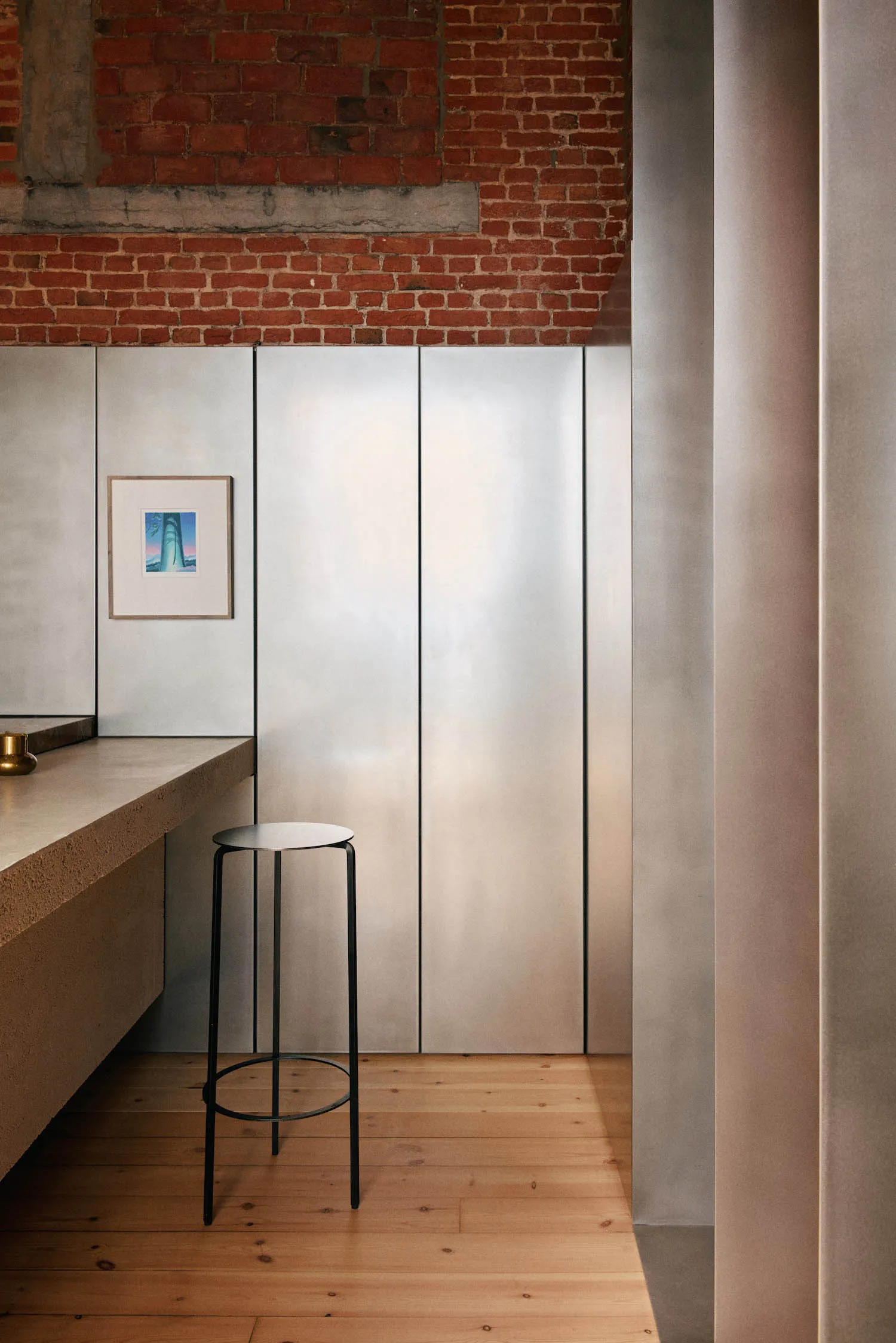 Modern industrial kitchen interior featuring a light concrete countertop, black metal bar stool, large stainless steel cabinetry, exposed red brick wall, and light wood flooring.