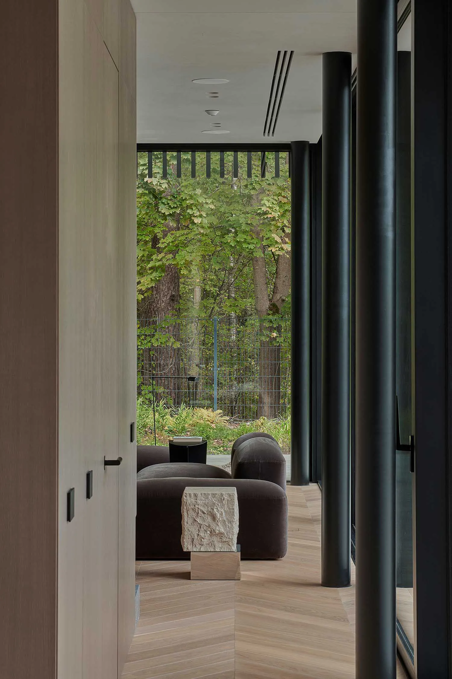 Modern living room interior with dark brown modular sofas, a raw stone ottoman on a wooden base, light oak herringbone flooring, black steel columns, and a large glass wall overlooking a lush green forest.