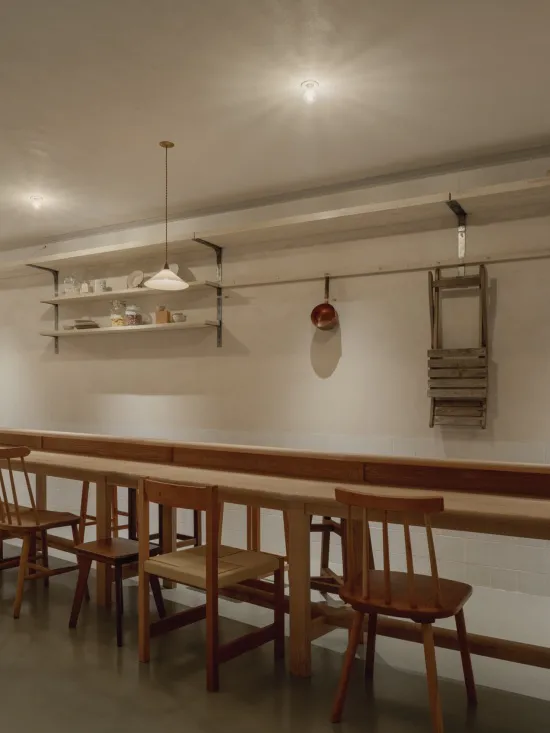 長型木製共享餐桌搭配橡木椅,漂浮層板上擺放餐具,懸掛銅鍋與木梯靠在白色磁磚牆上。