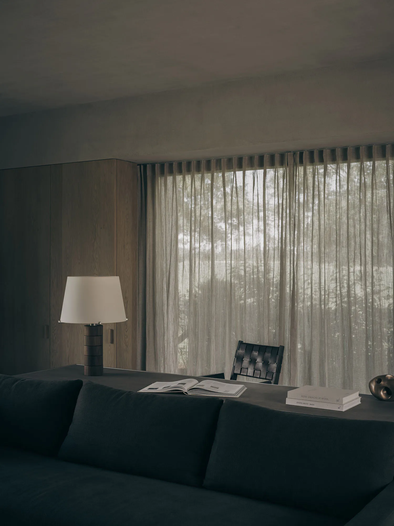 Modern minimalist living room interior featuring a dark teal sofa in the foreground, a wooden table with an open book and table lamp, a woven black leather chair, floor-to-ceiling sheer curtains, and warm wood paneling under soft natural light.