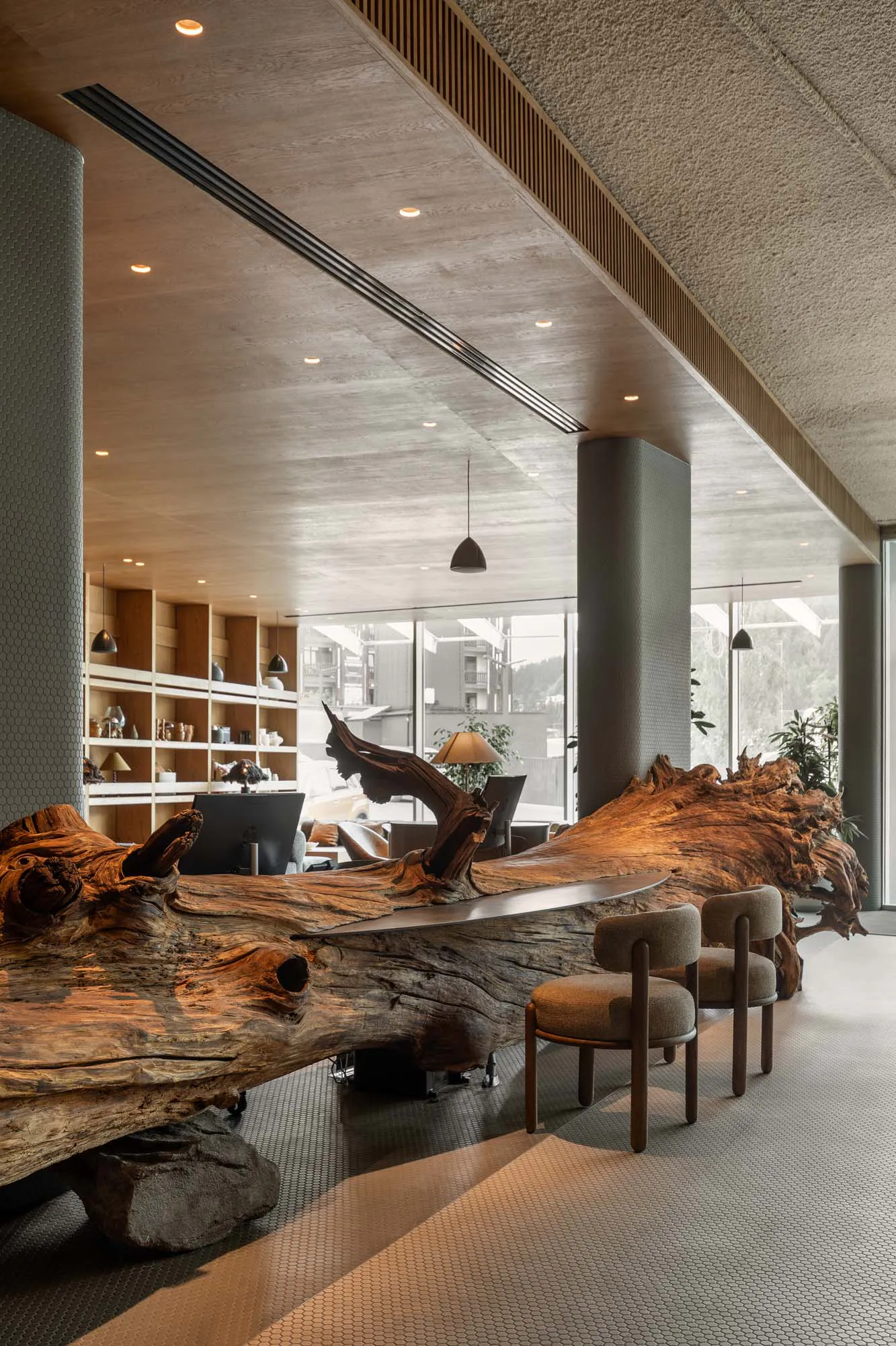 Modern lobby interior featuring an enormous live-edge wood reception desk made from a massive tree trunk, paired with upholstered chairs and concrete architectural elements.
