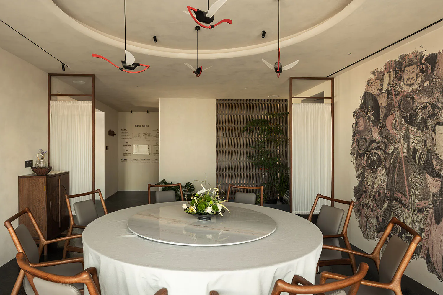 Modern Chinese-style dining room with a large round marble lazy Susan table, wooden dining chairs with grey upholstery, dramatic Tang dynasty mural on the right wall, and artistic red and white ceiling fans hanging from a curved ceiling.