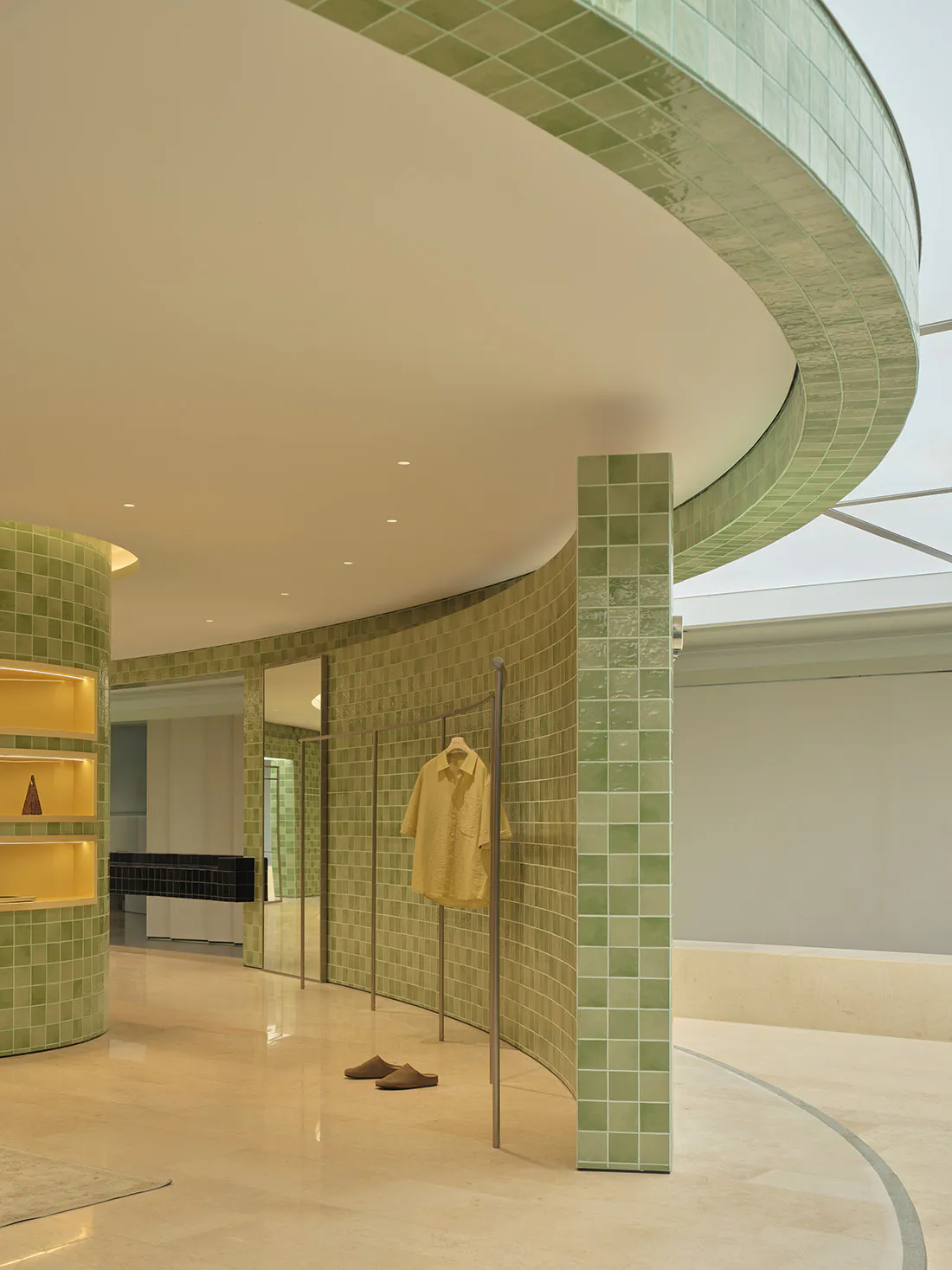 Modern boutique interior featuring curved walls covered in glossy green ceramic tiles, a minimalist clothing rack with a yellow shirt, and a cylindrical shelving unit with warm backlighting.