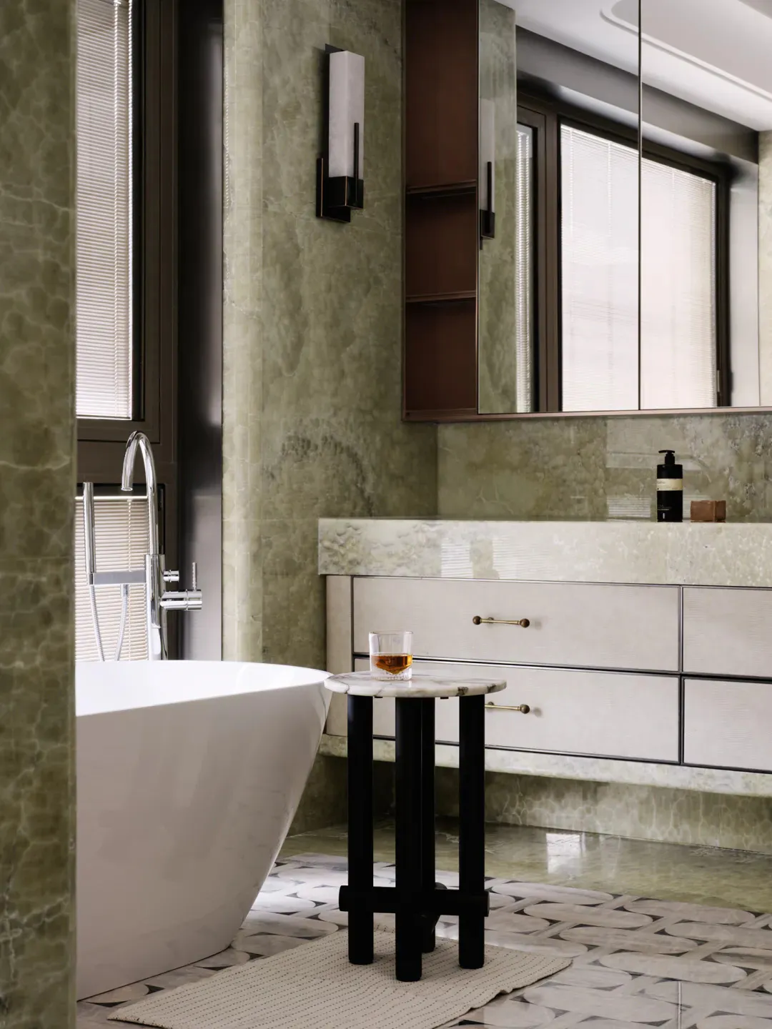 Luxurious bathroom interior with green marble walls, white freestanding bathtub, floating vanity with drawers, large mirror, and a black and white marble side table holding a glass of amber liquid.